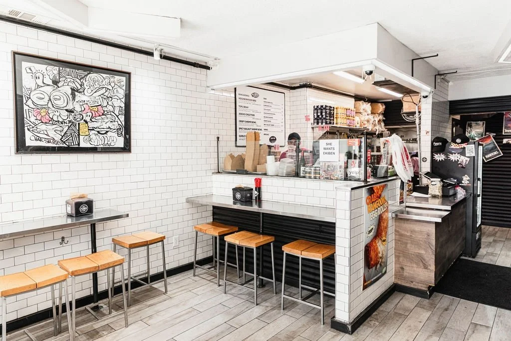 Interior of a small takeout restaurant or cafe with white tiled walls, a counter with a glass display, and stools for seating. There are signs, a menu, and caps on display.