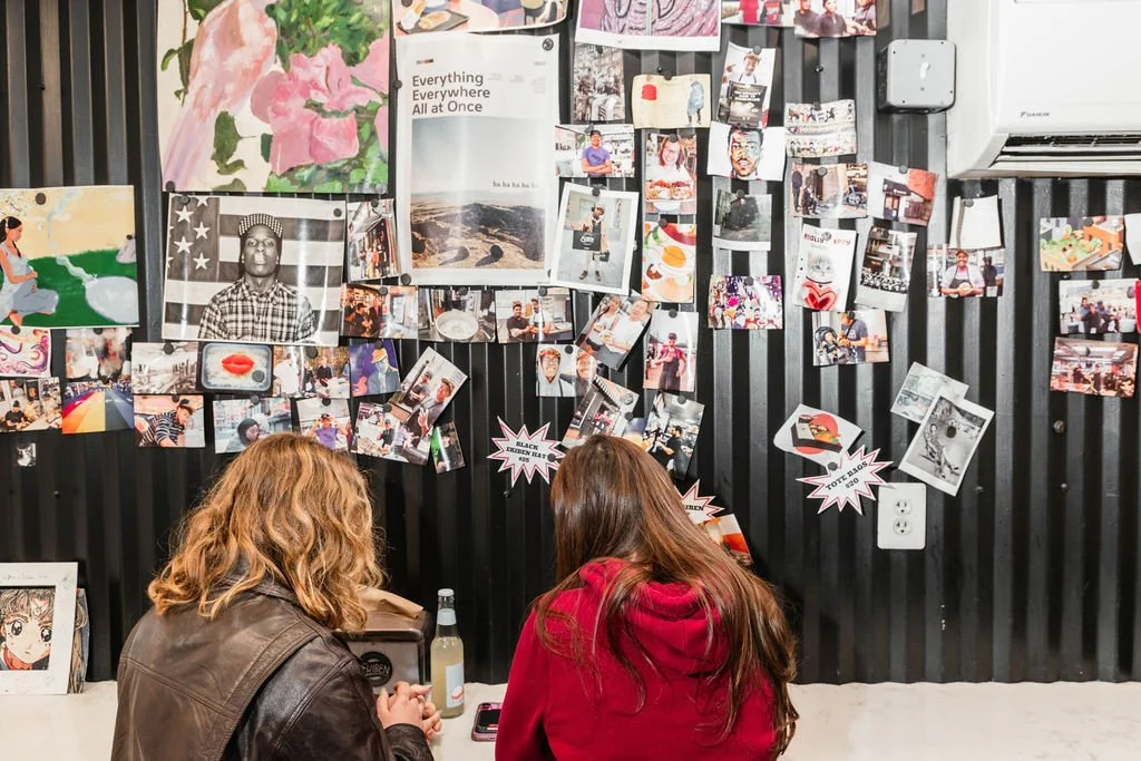 Two women with brown hair sitting at a table with drinks, facing a black wall decorated with various photos, artwork, and paper notes.