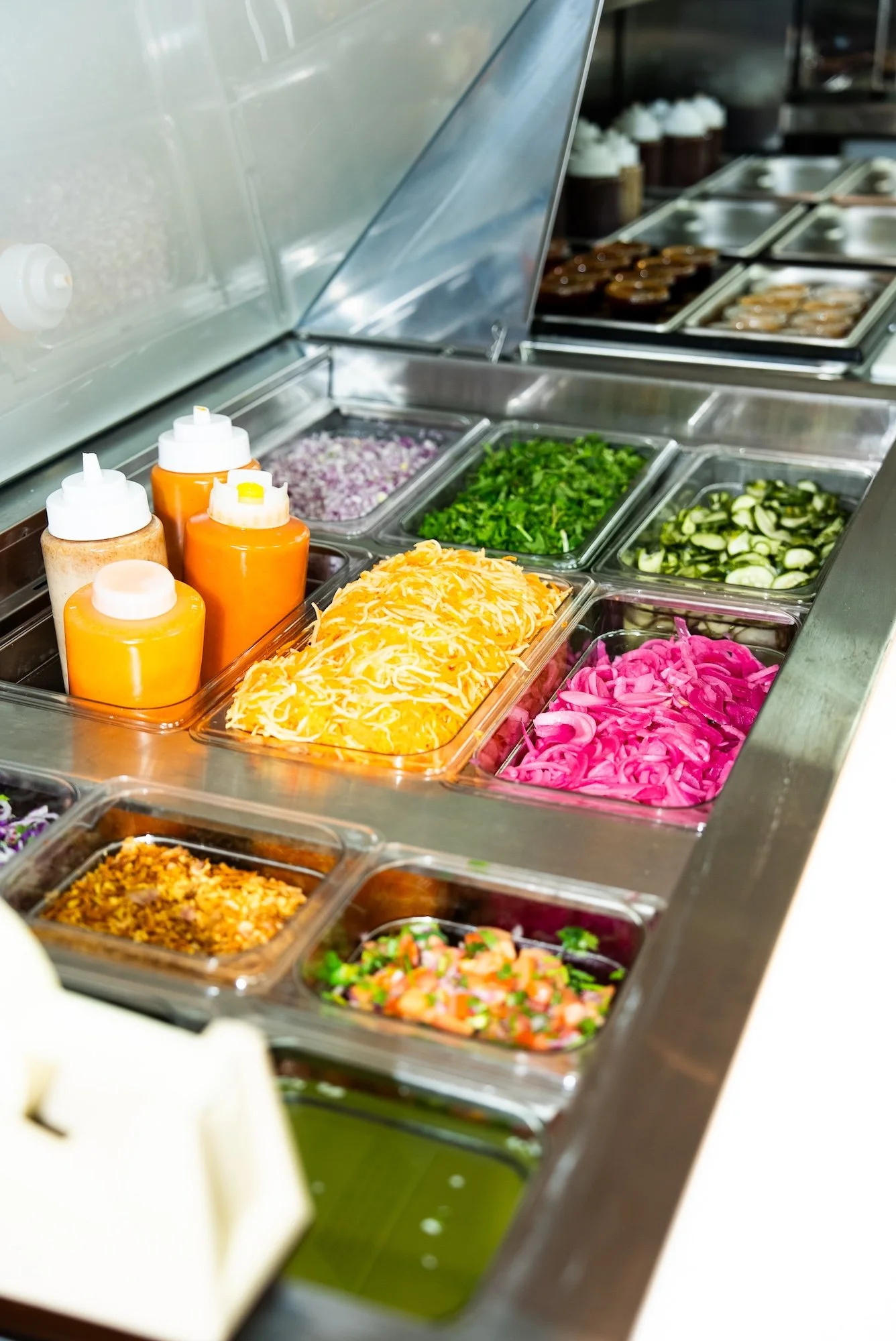 A buffet line with various toppings and condiments, including sliced pickles, shredded cheese, chopped onions, chopped cilantro, pink pickled onions, and sauces in squeeze bottles, with desserts like cupcakes and cookies visible in the background.