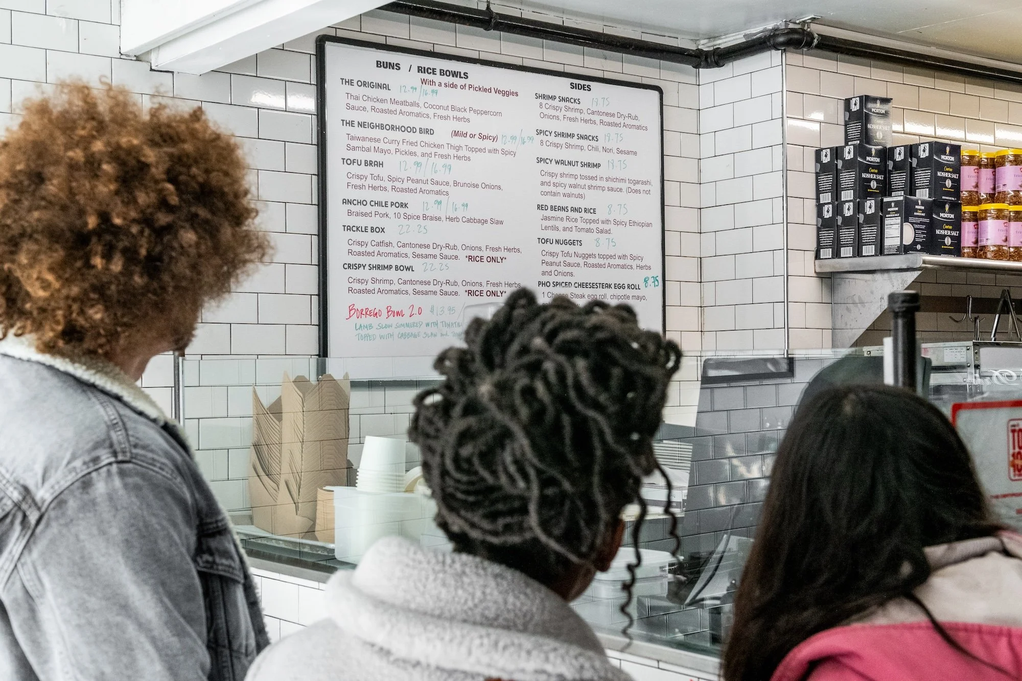 People standing at a counter inside a restaurant or food shop, with a menu board on the white tiled wall behind them listing various dishes and prices.