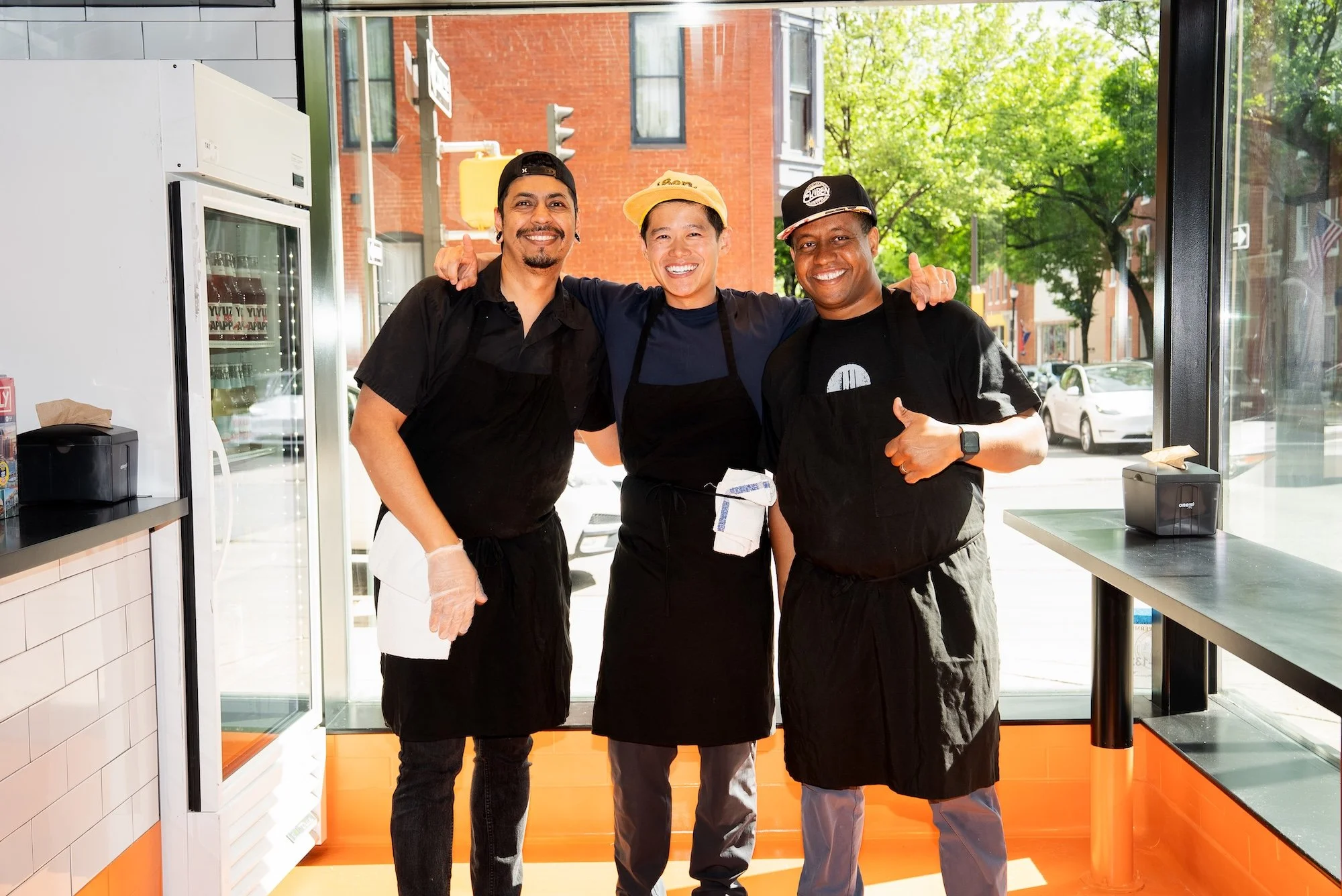 Three men standing in a restaurant, smiling and posing for the camera, wearing aprons and hats, with a window showing trees and a brick building outside.
