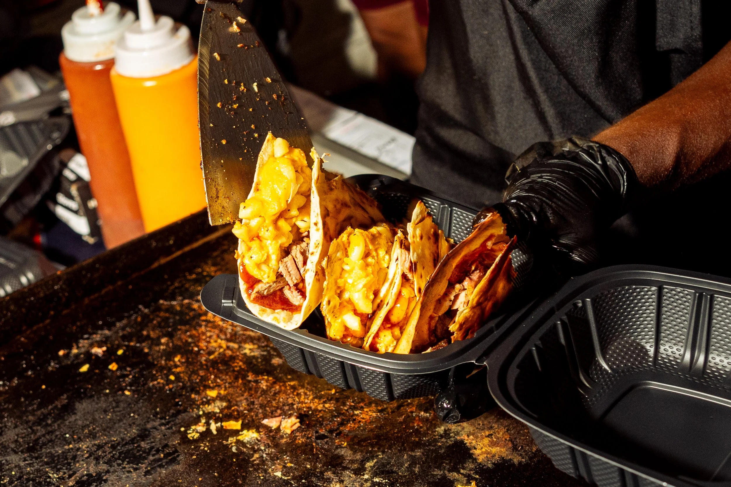 A person wearing black gloves and a dark shirt preparing tacos with scrambled eggs, meat, and cheese inside soft tortillas on a black countertop.