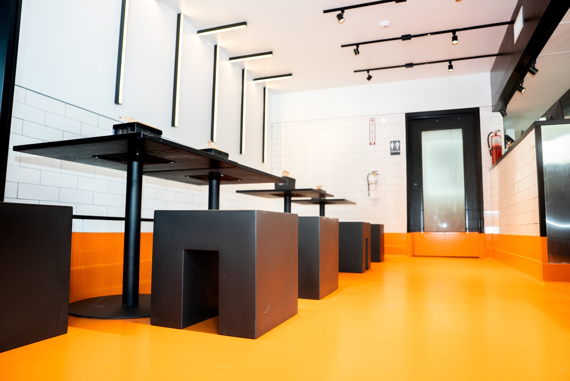 Empty modern dining area with black tables and stools, orange flooring, white brick wall, and black details on the ceiling and doorframe.