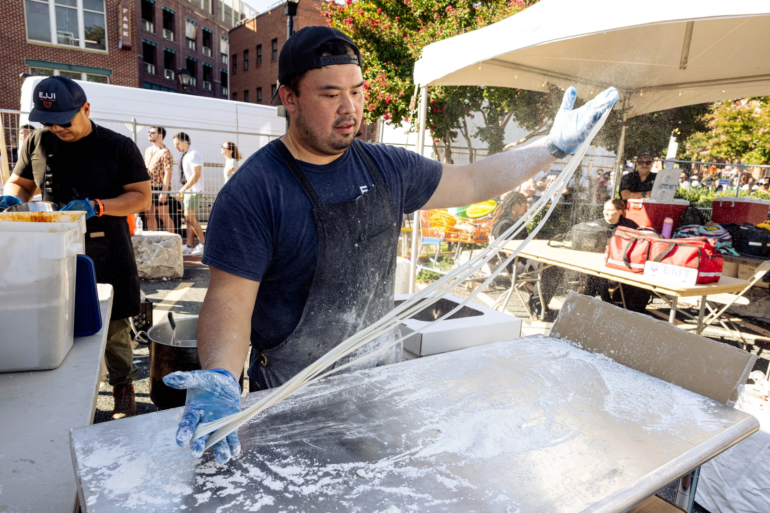 A man wearing a black cap, blue shirt, and gloves is preparing food outdoors. He is working at a table with various items, and there is a spray of water or dust in the air as he works. Several people are in the background, along with trees, a fence, and a white tent, indicating an outdoor event or market.