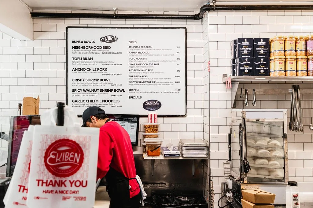 Interior of a restaurant with a menu board on white tiled wall, employee in red uniform working at the counter, and shelves with canned goods and bakery items.