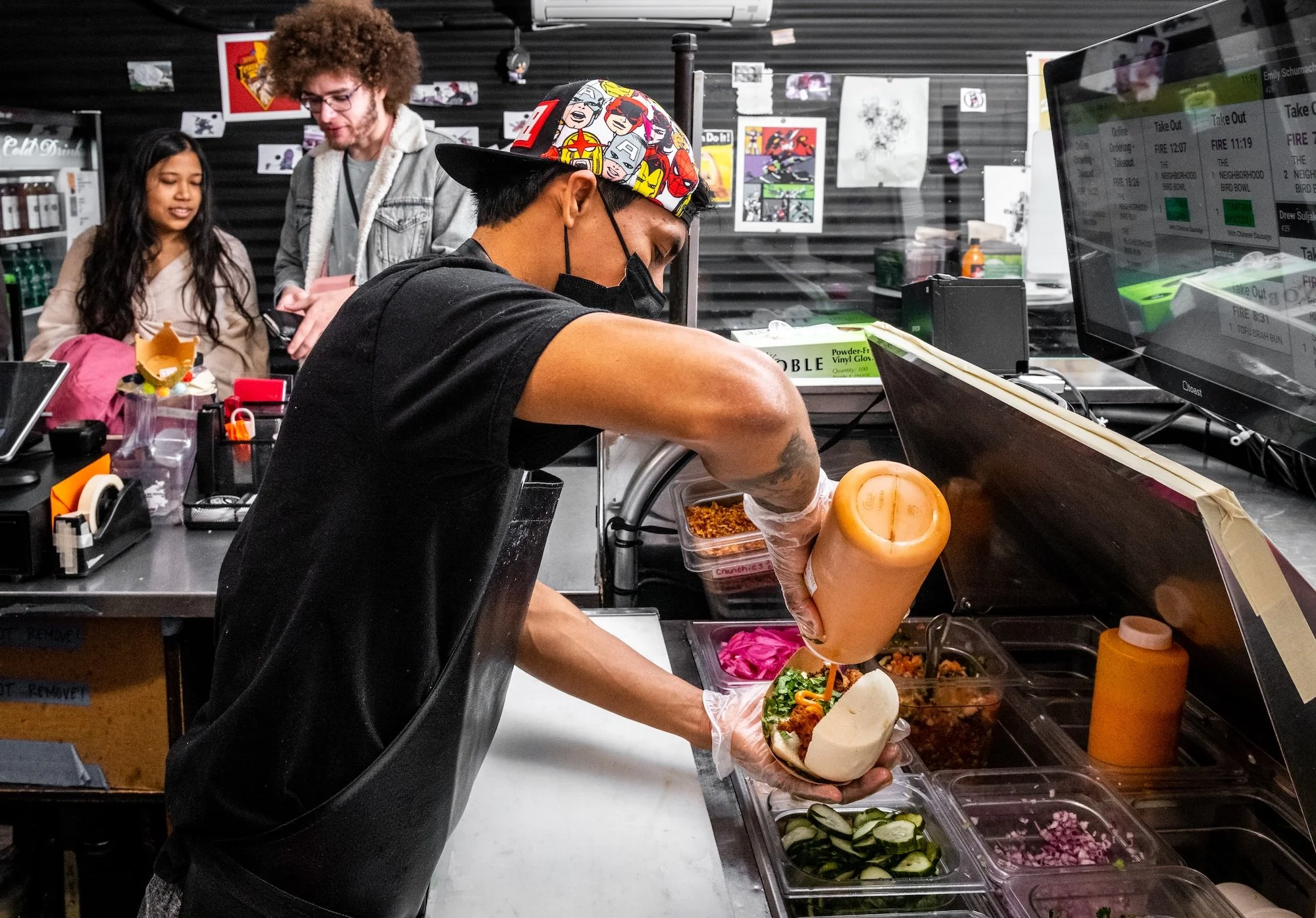 A person wearing a colorful cap, black mask, and black shirt is preparing food at a deli or restaurant counter, holding a prepared sandwich or wrap, with fresh vegetables in containers in front.