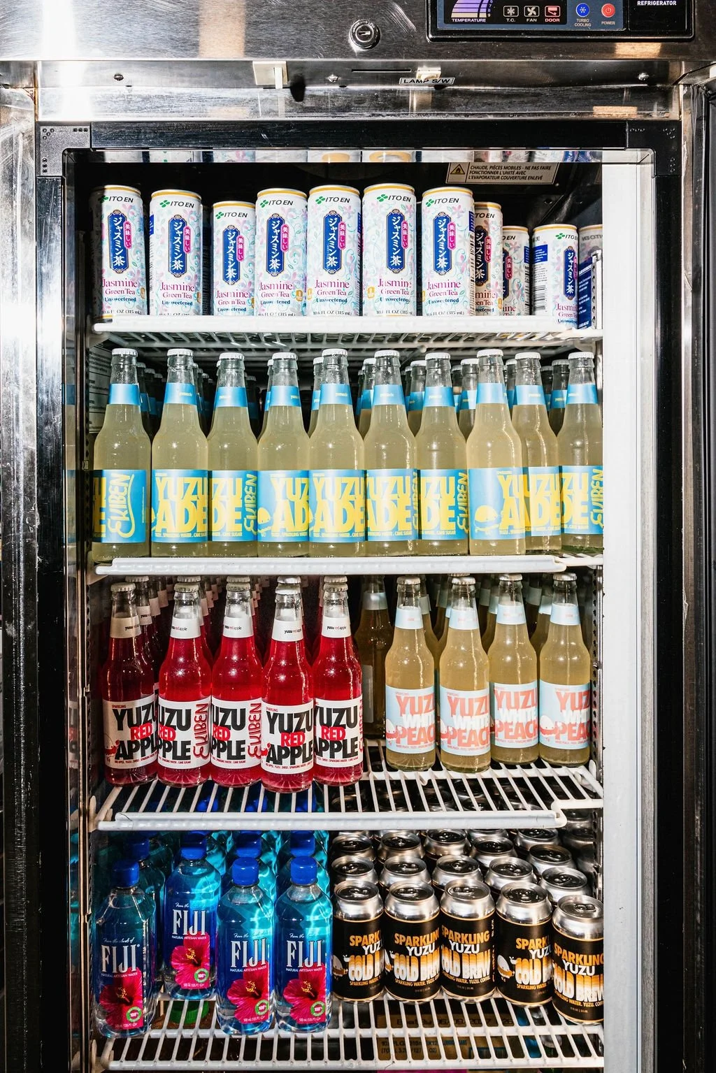 A refrigerator stocked with various bottled beverages, including jasmine green tea at the top, Yuz Adesen soda in the middle, and Fiji water and Sparkling Yuzu cans at the bottom.
