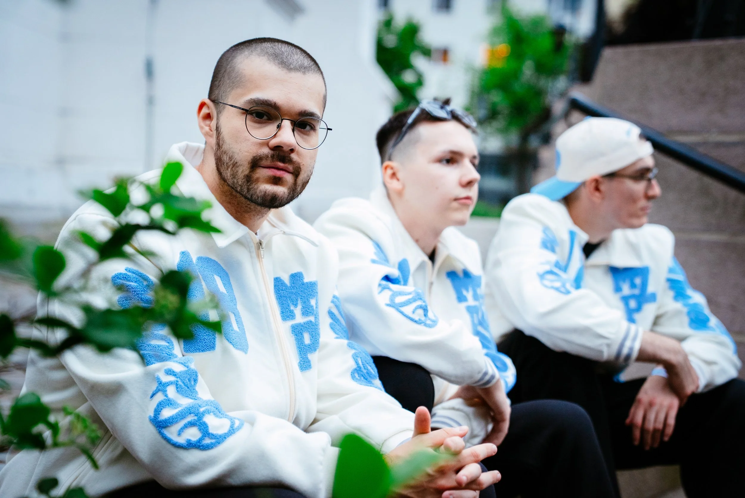 Tre unge menn i hvite jakker med blå logo, sitter på rekke utendørs, en av dem ser direkte på kamera, mens de andre ser bort. Bakgrunn med grønne planter og en trapp.