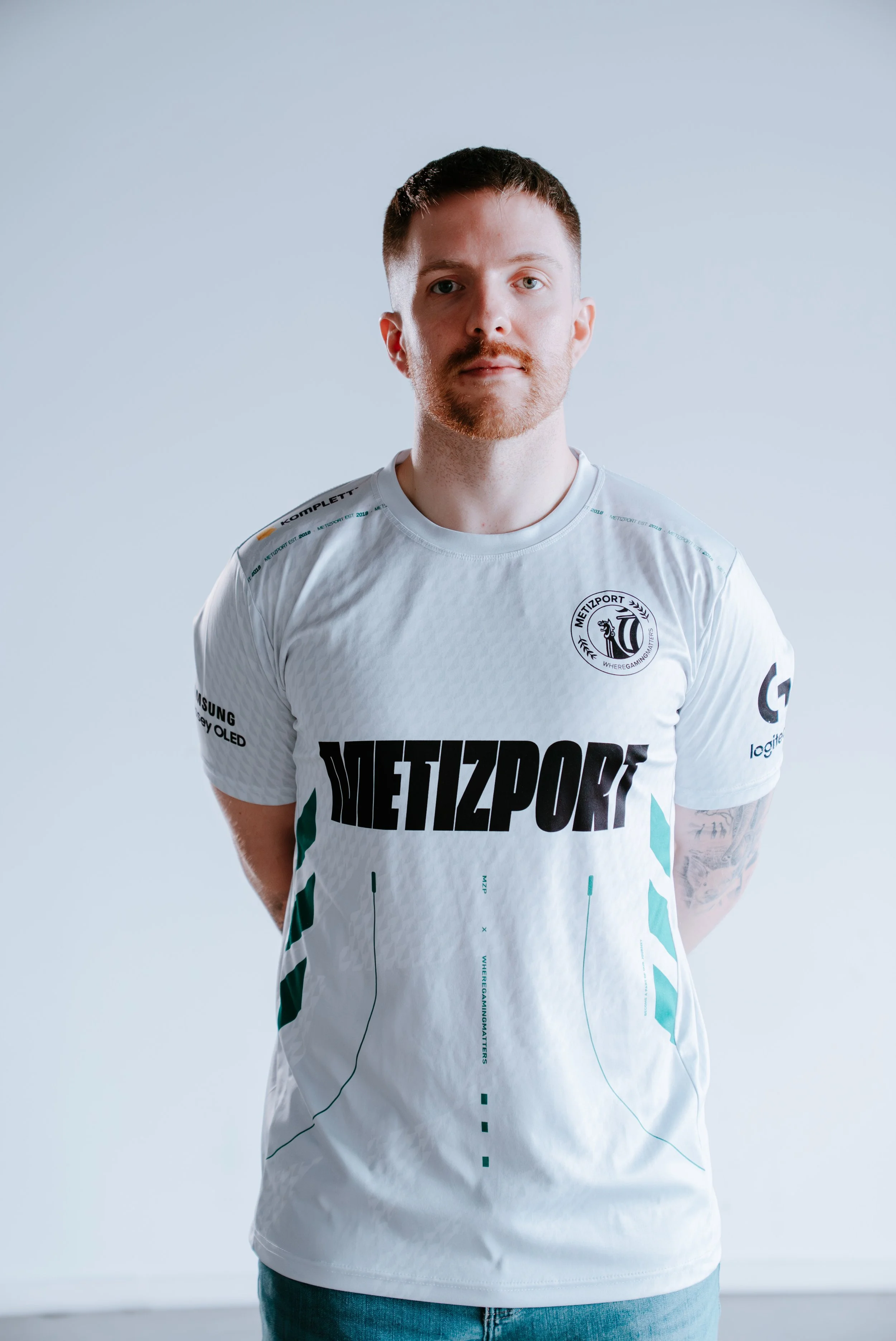 En mann med kort brunt hår og skjegg, iført en hvit esport-trøye med logoer og teksten 'METZPORT', står foran en lys grå bakgrunn.