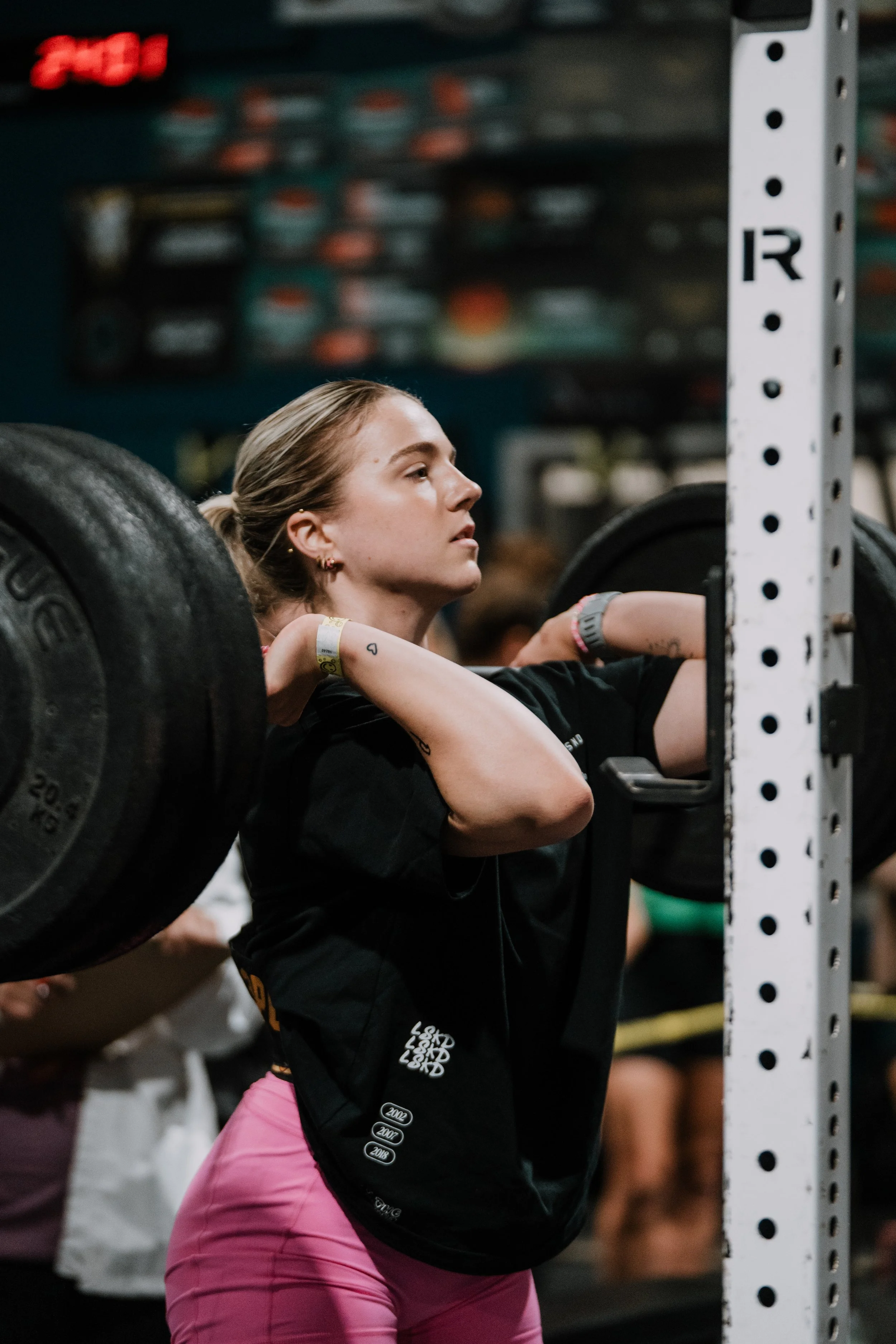 Young woman lifting a barbell with weight plates in a gym, focused, wearing pink leggings and a black shirt, with a tattoo on her arm.