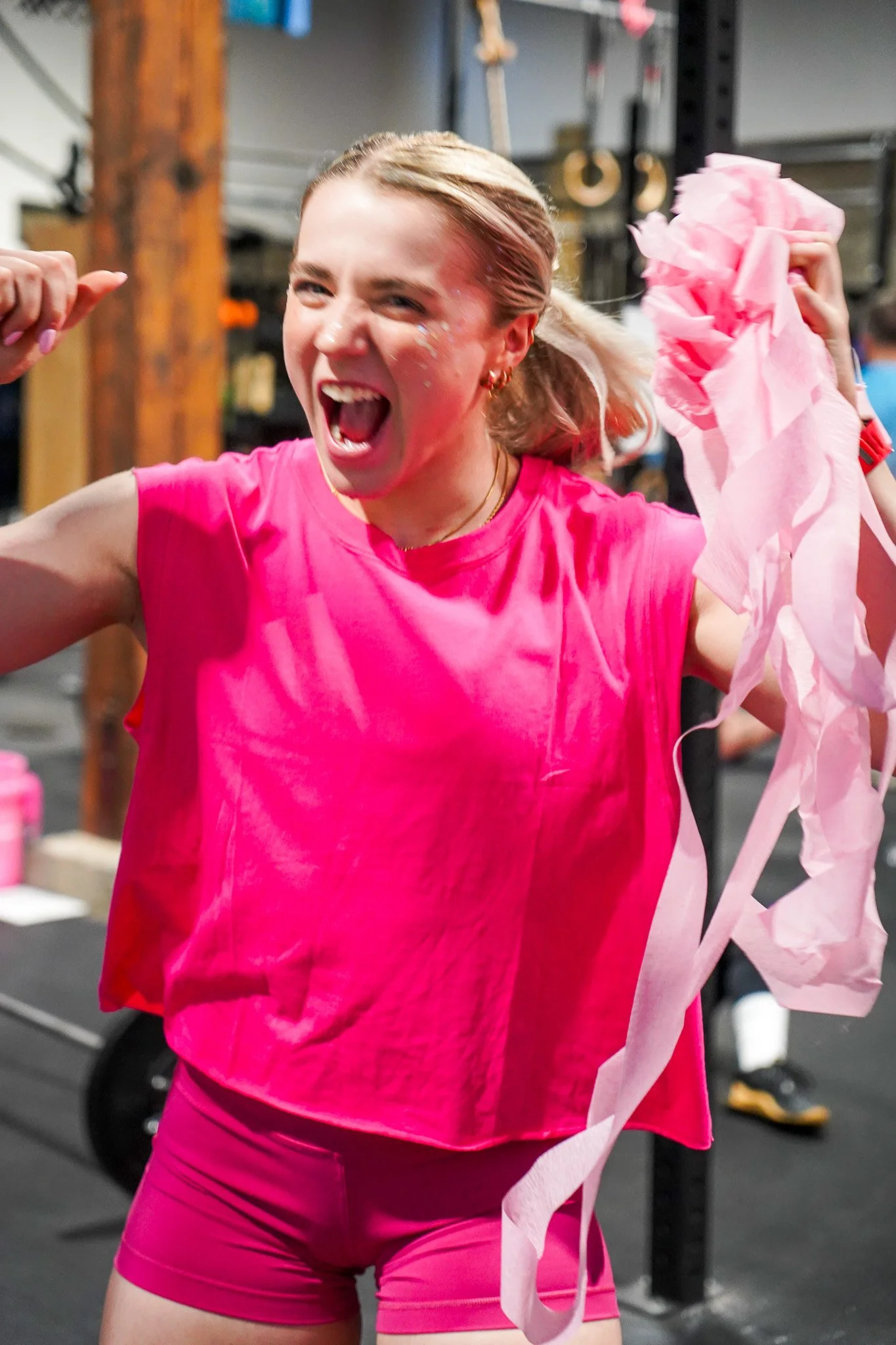 A woman in a bright pink shirt and shorts, celebrating with a pink ribbon in a gym, smiling and showing a flexed arm.