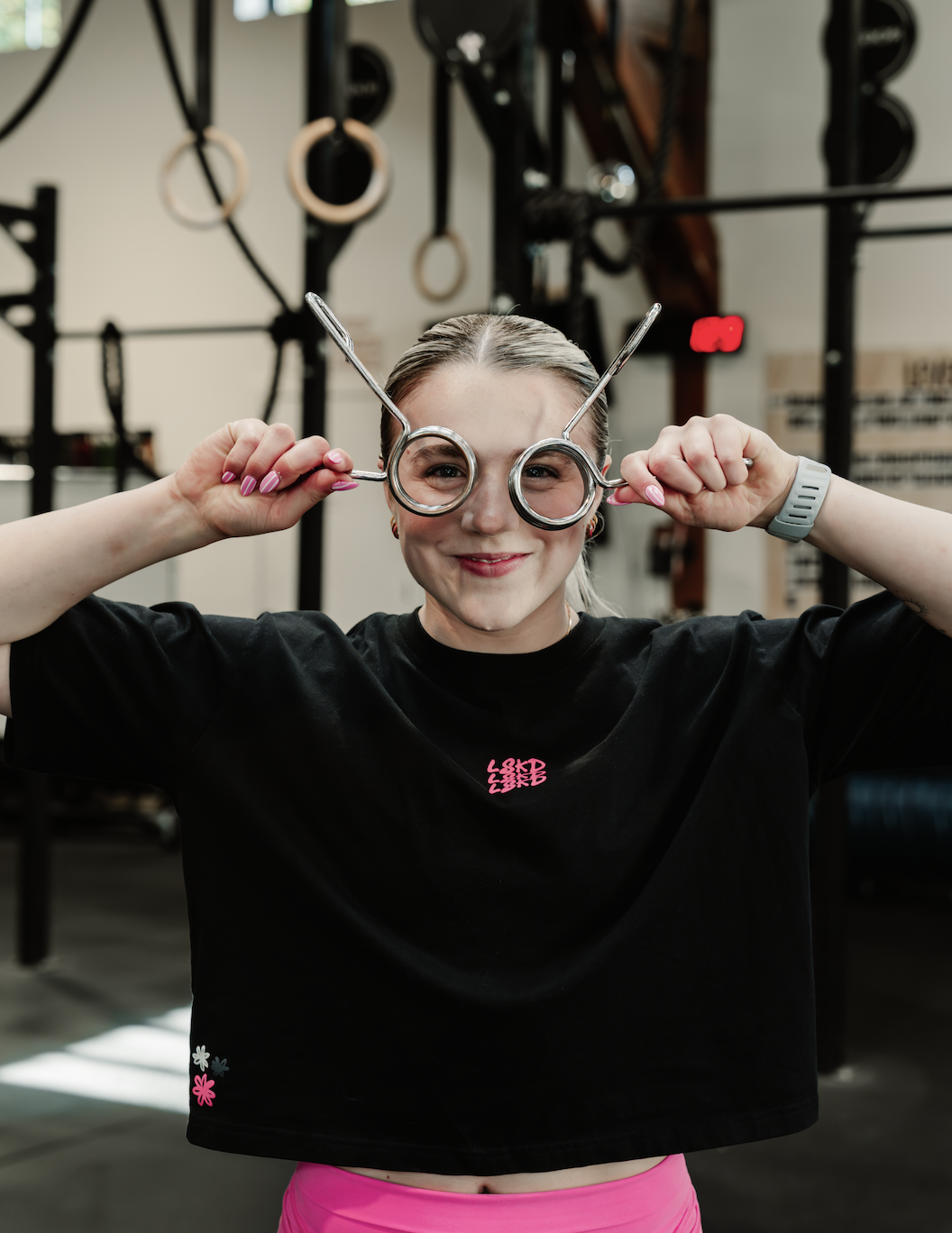 A young woman in a gym holding gymnastic rings around her eyes and smiling.