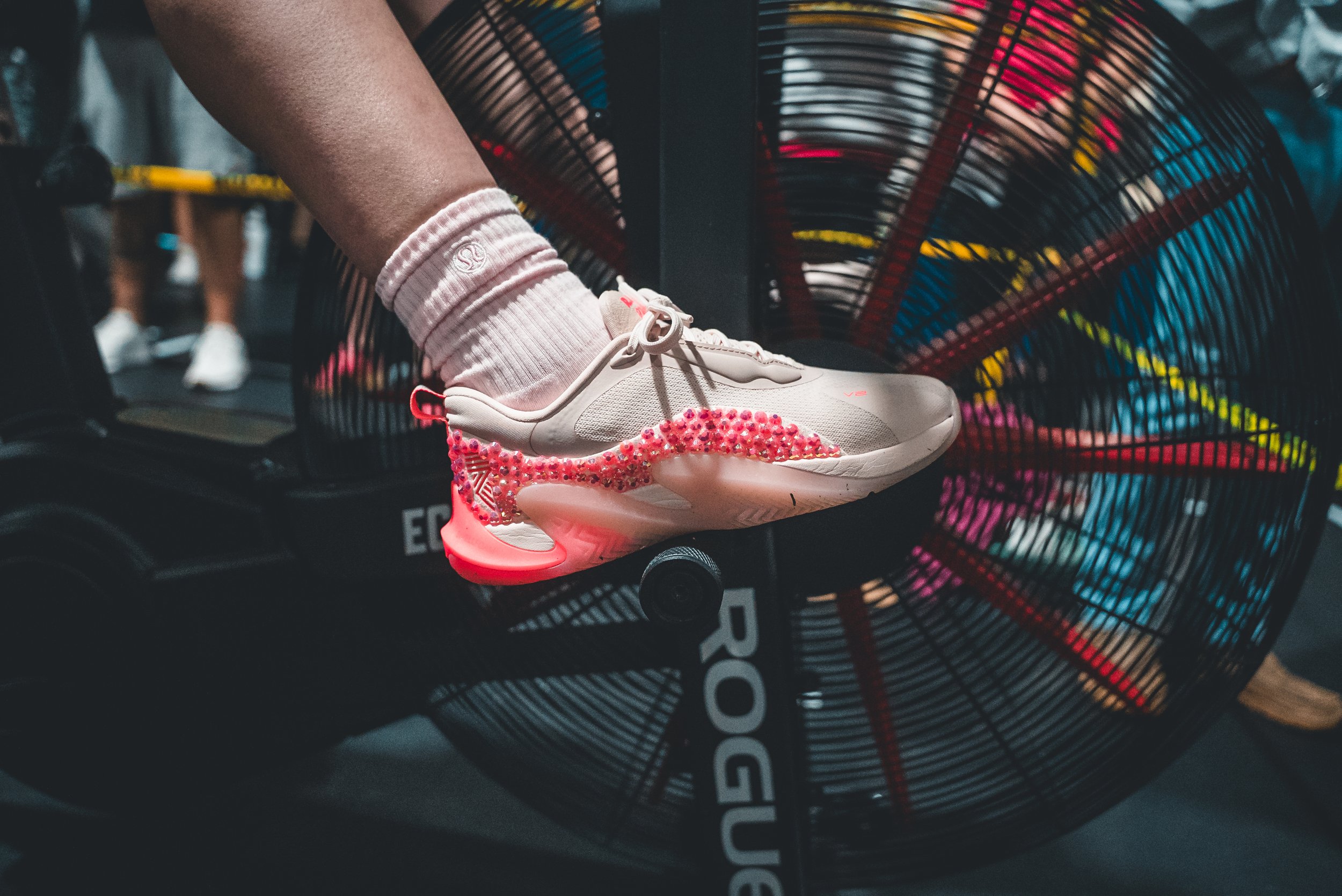 A person wearing white athletic shoes and pink socks stepping on a treadmill, with a fan behind them on a gym floor.