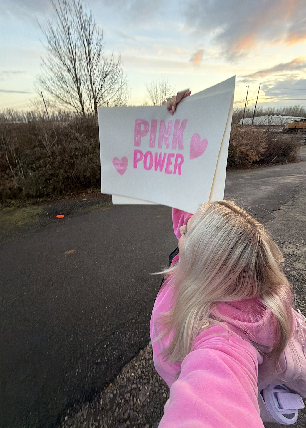 A person holding a sign that reads "Pink Power" with pink hearts, wearing a pink hoodie, standing outdoors during sunset with leafless trees and a cloudy sky in the background.