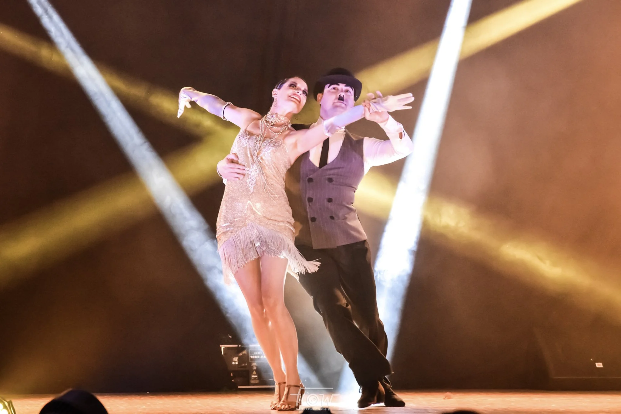Un couple danse sur scène avec un éclairage de scène dramatique, la femme porte une robe de danse en or et le homme une veste marron et un chapeau noir.