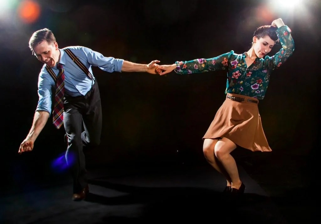 Un homme et une femme dansent ensemble sur scène, l'homme tend la main à la femme tandis qu'elle semble hésiter, dans un style de danse rétro ou jazz, avec un éclairage dramatique.