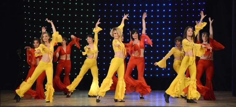 Groupe de femmes en costumes colorés de danse sur scène, en position de danse fluide, avec un fond lumineux.