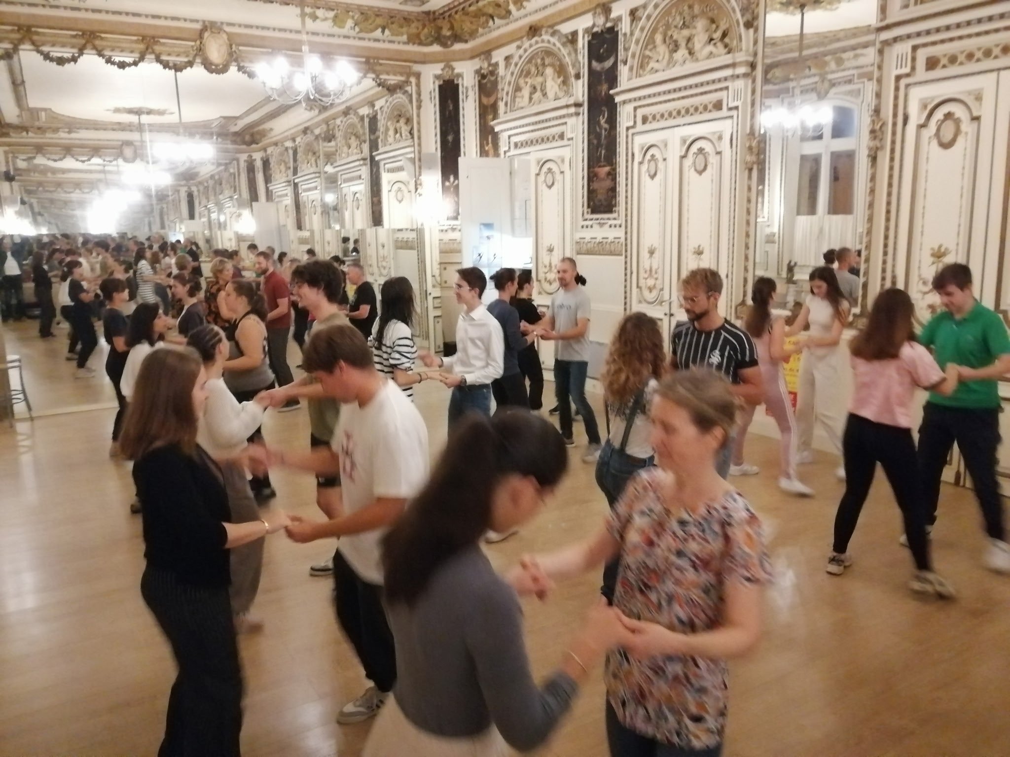Groupe de personnes dansant lors d'une soirée dans une salle ornée de décorations classiques et de lustres chandeliers.
