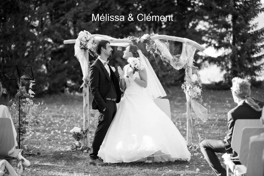 Un couple de mariés échangeant leurs vœux lors d'une cérémonie de mariage en plein air, sous un arc floral, avec des invités assis autour.