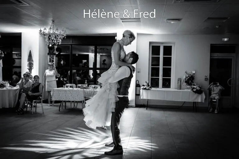 Un couple danse lors d'un mariage, la femme en robe de mariée et l'homme en costume, dans une salle décorée pour un événement, avec des invités assis à des tables en arrière-plan.