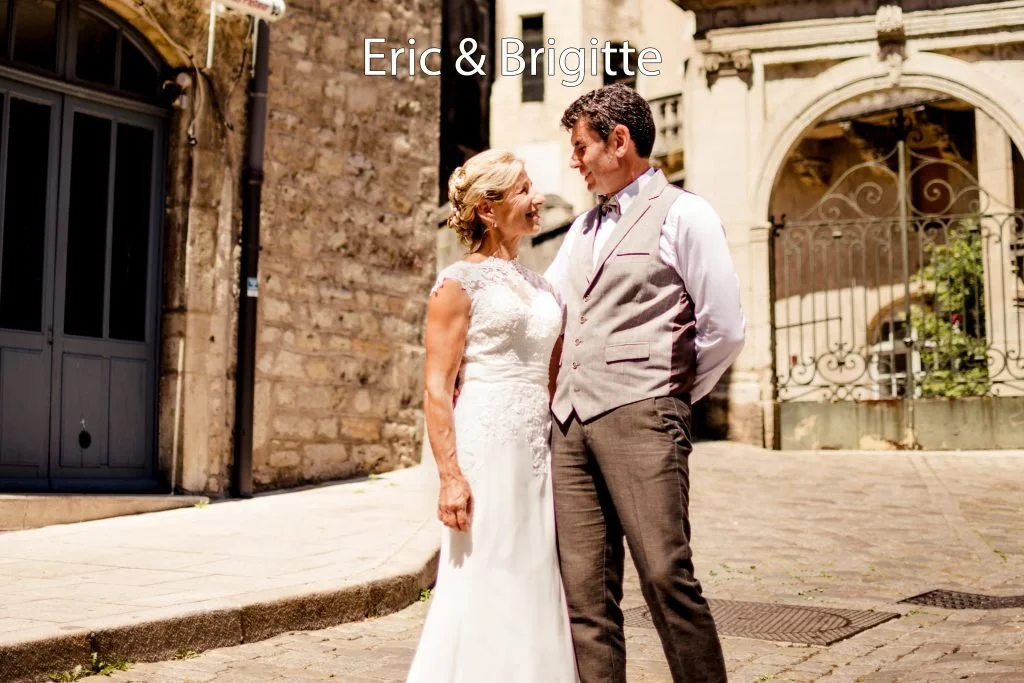 Un couple, une femme en robe de mariage et un homme en costume, sourient et regardent l'un l'autre dans une rue pavée avec un fond de bâtiments anciens en pierre.