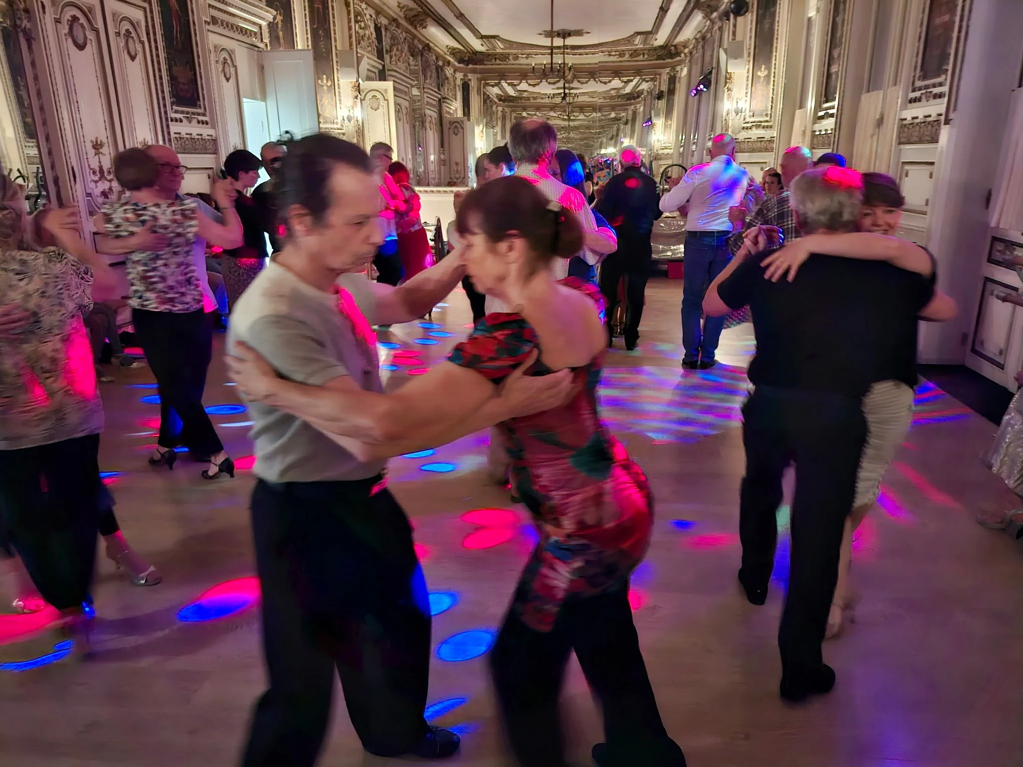 Des personnes dansant dans une salle ornée de décorations classiques, sous des lumières colorées, lors d'une fête ou d'un événement social.