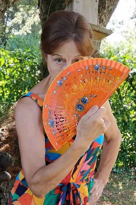 Une femme âgée tenant un éventail orange en cachetant une partie de son visage en plein air avec un fond de feuillage vert et d'arbres.