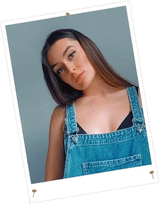 Une jeune femme aux cheveux longs et bruns pose en portant une salopette en jean noire et un haut noir, avec un fond gris.