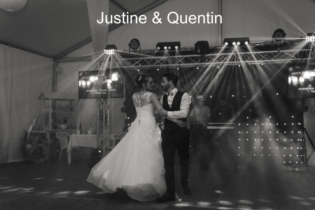 Un couple danse lors d'une réception de mariage sous un chapiteau, lumière de scène et décoration en arrière-plan, en noir et blanc.