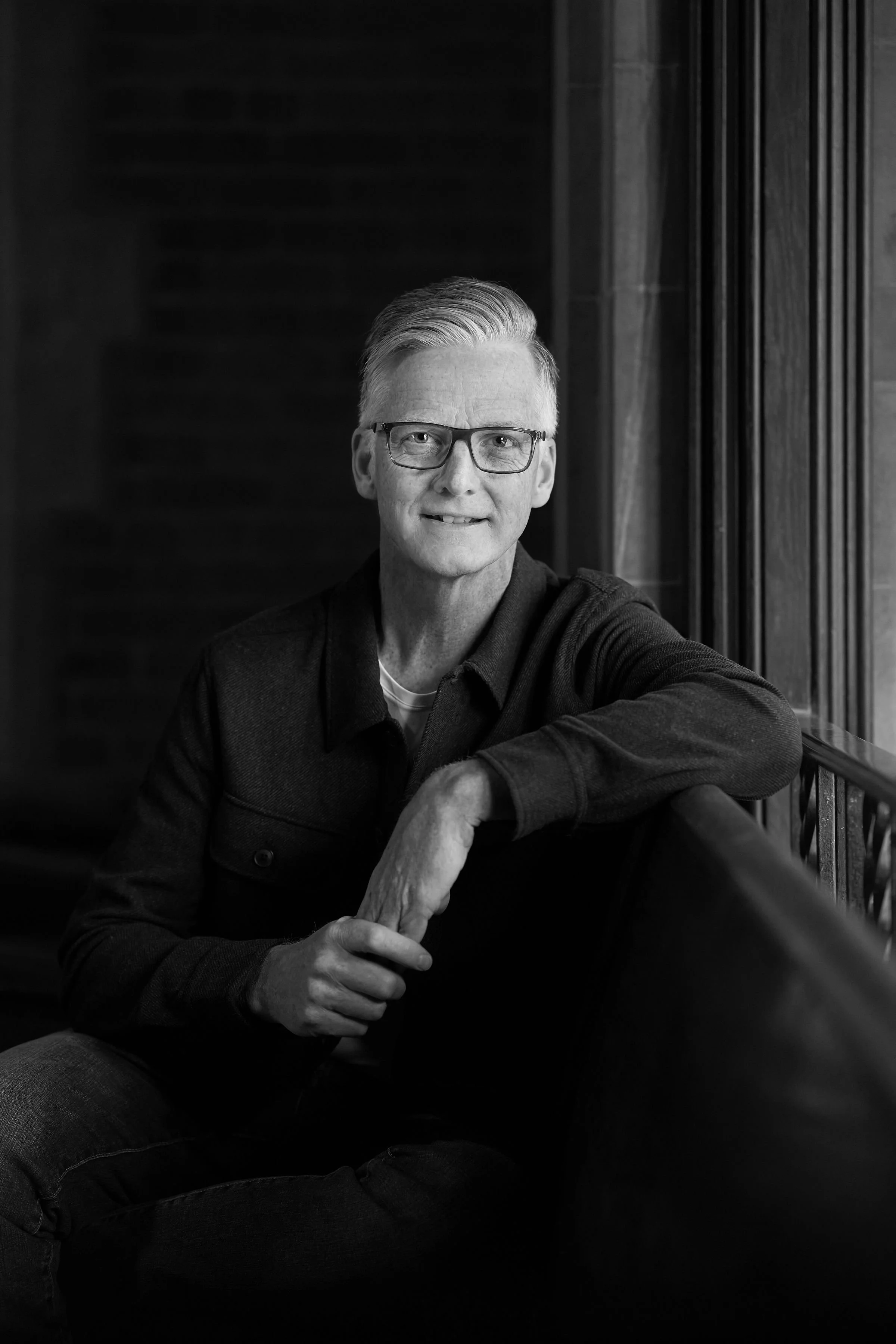 Black and white photo of a middle-aged man with glasses and neatly combed back hair, smiling slightly, sitting indoors next to a wooden wall with vertical paneling, resting his arm on his knee.