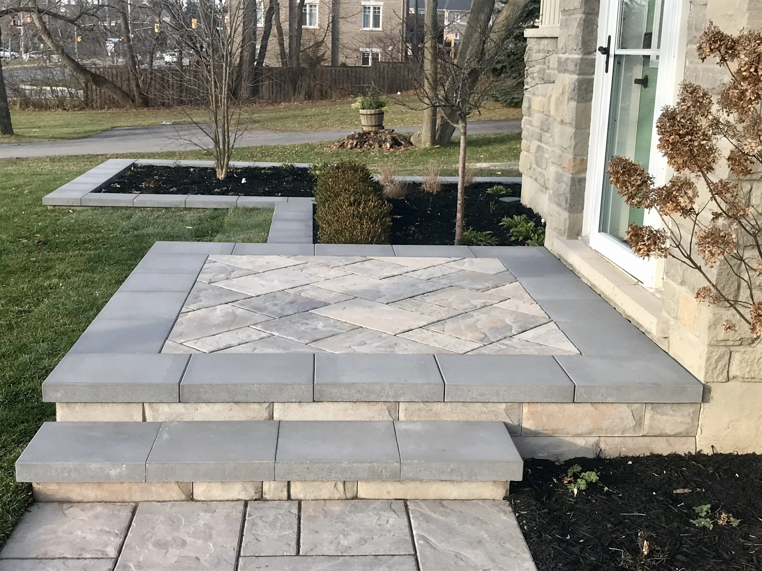 A backyard patio with stone steps leading down to a grassy area. There are plants and bushes along the side of a house with a stone facade, leading to a sidewalk and a park with trees and an apartment building in the background.