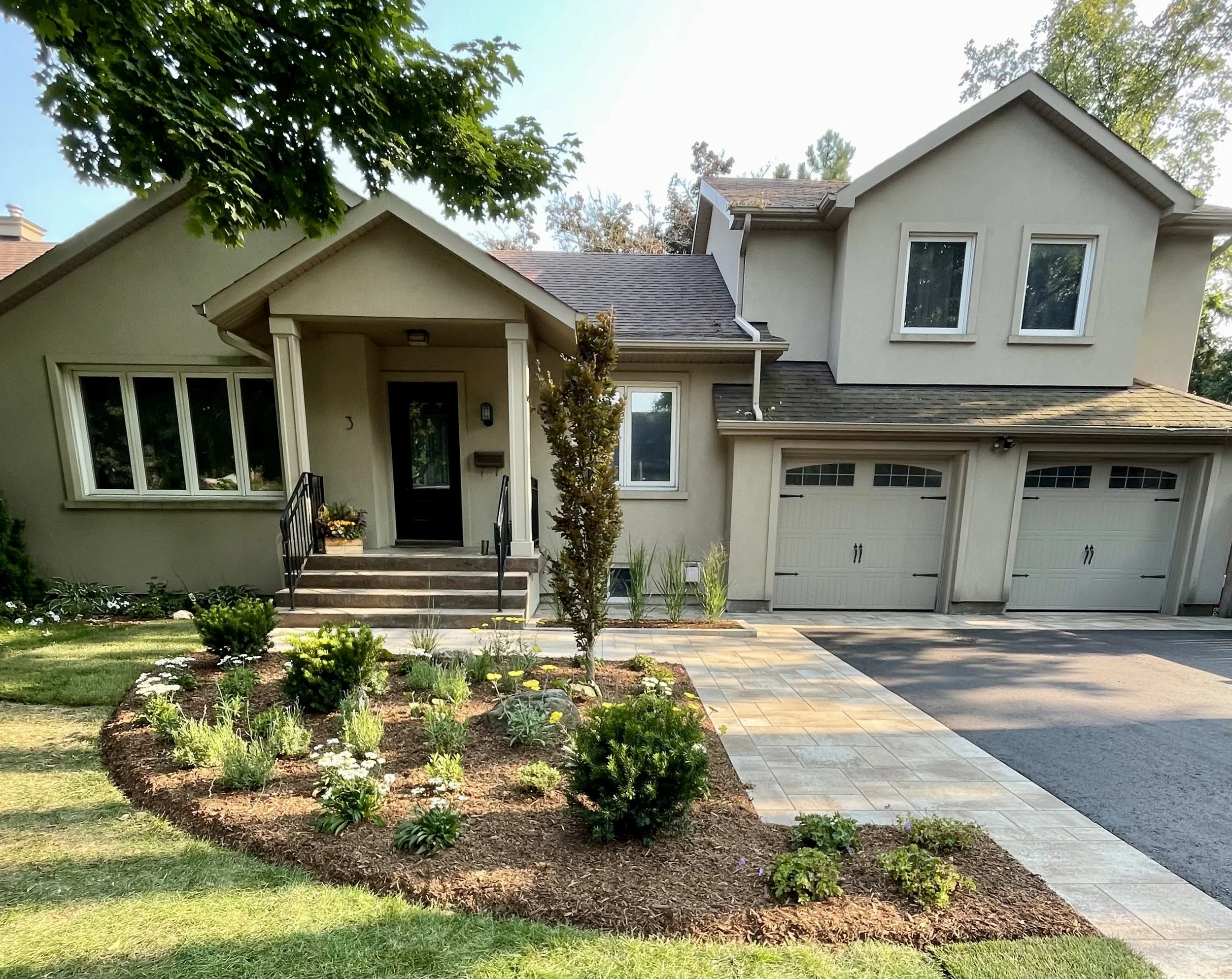 Front yard of a house with a landscaped garden, a stone walkway, and a driveway with garage doors. The house has a cream exterior with multiple windows and a covered front porch.