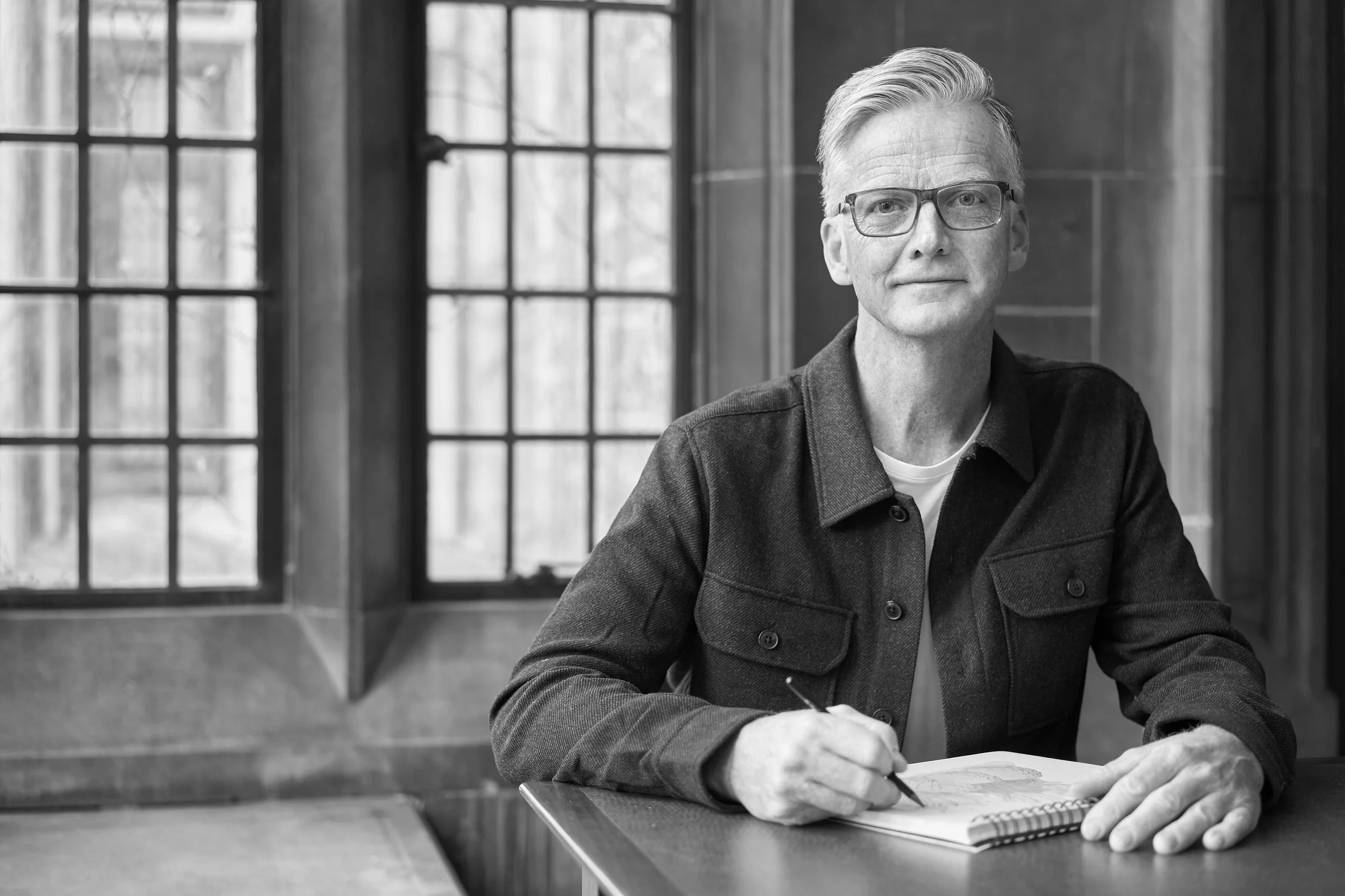 A black and white photo of an older man with glasses and gray hair sitting at a table in front of a large window. He is holding a pen and appears to be thinking or writing in a notebook.