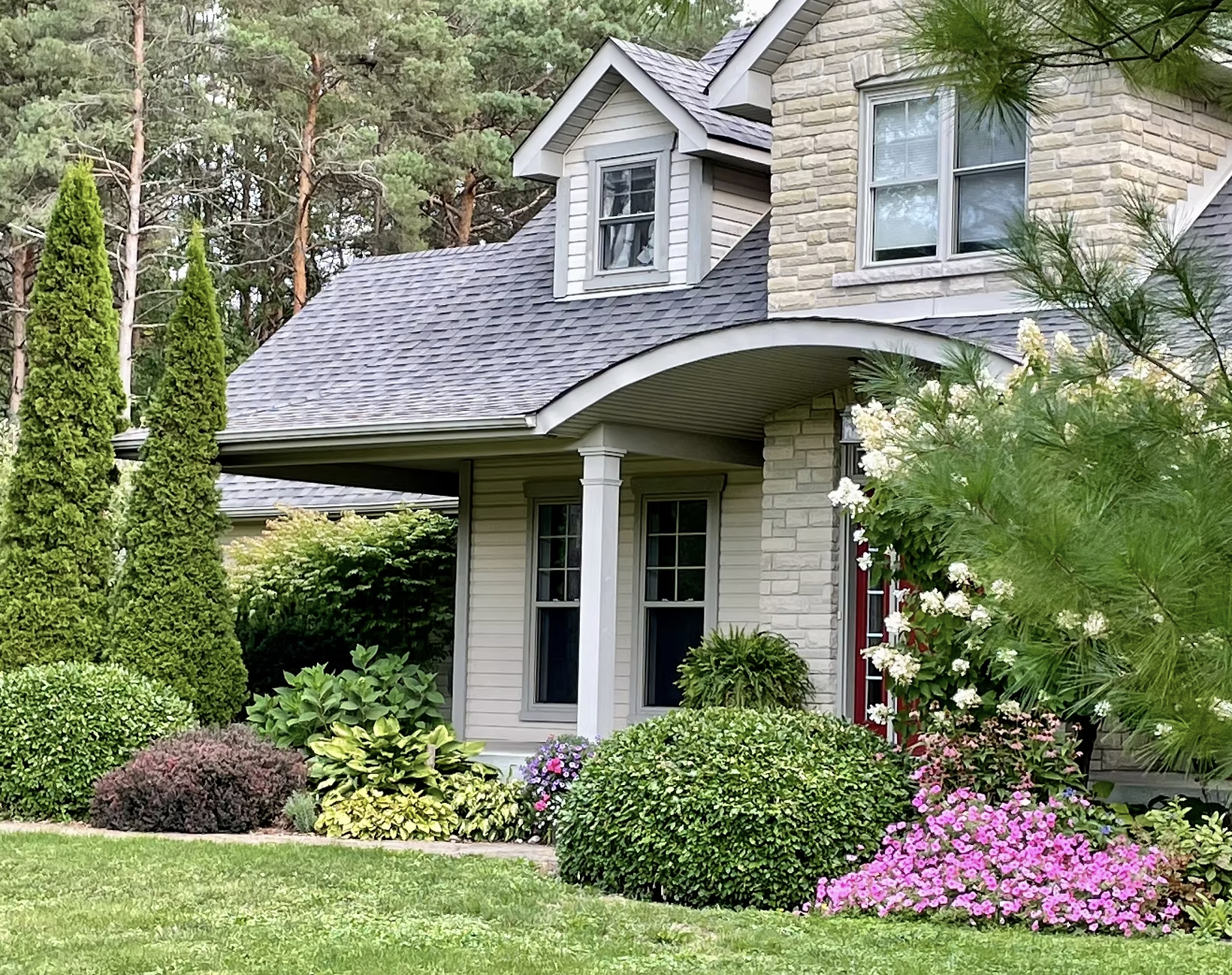 A cozy house with a pink brick exterior, surrounded by lush green lawn and vibrant flowering bushes, with tall trees in the background.