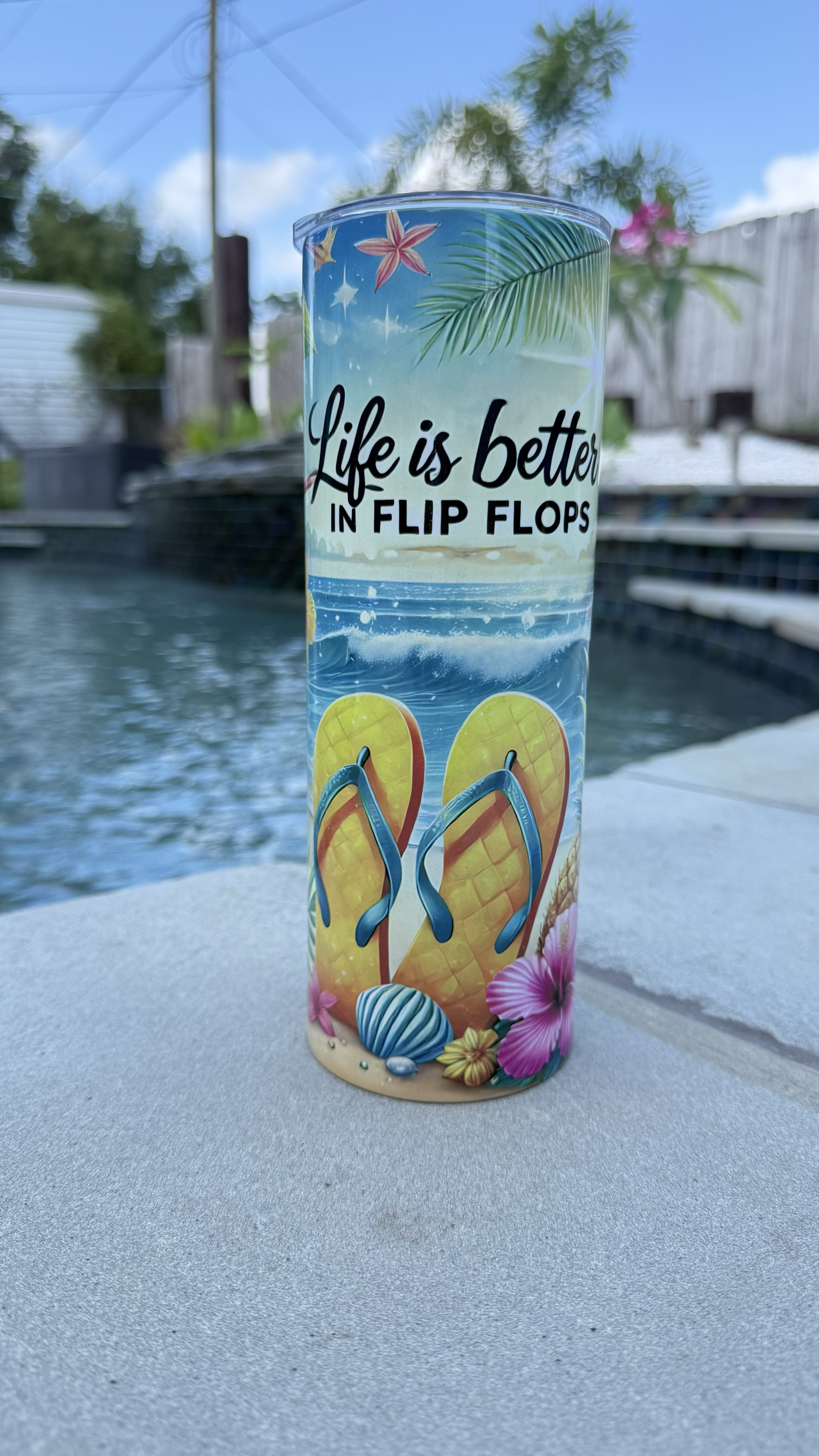 A colorful tumbler with a tropical beach scene, including flip flops, seashells, and flowers, with the phrase "Life is better in flip flops" written on it. It is set outdoors near a pool with a cloudy sky and palm trees in the background.