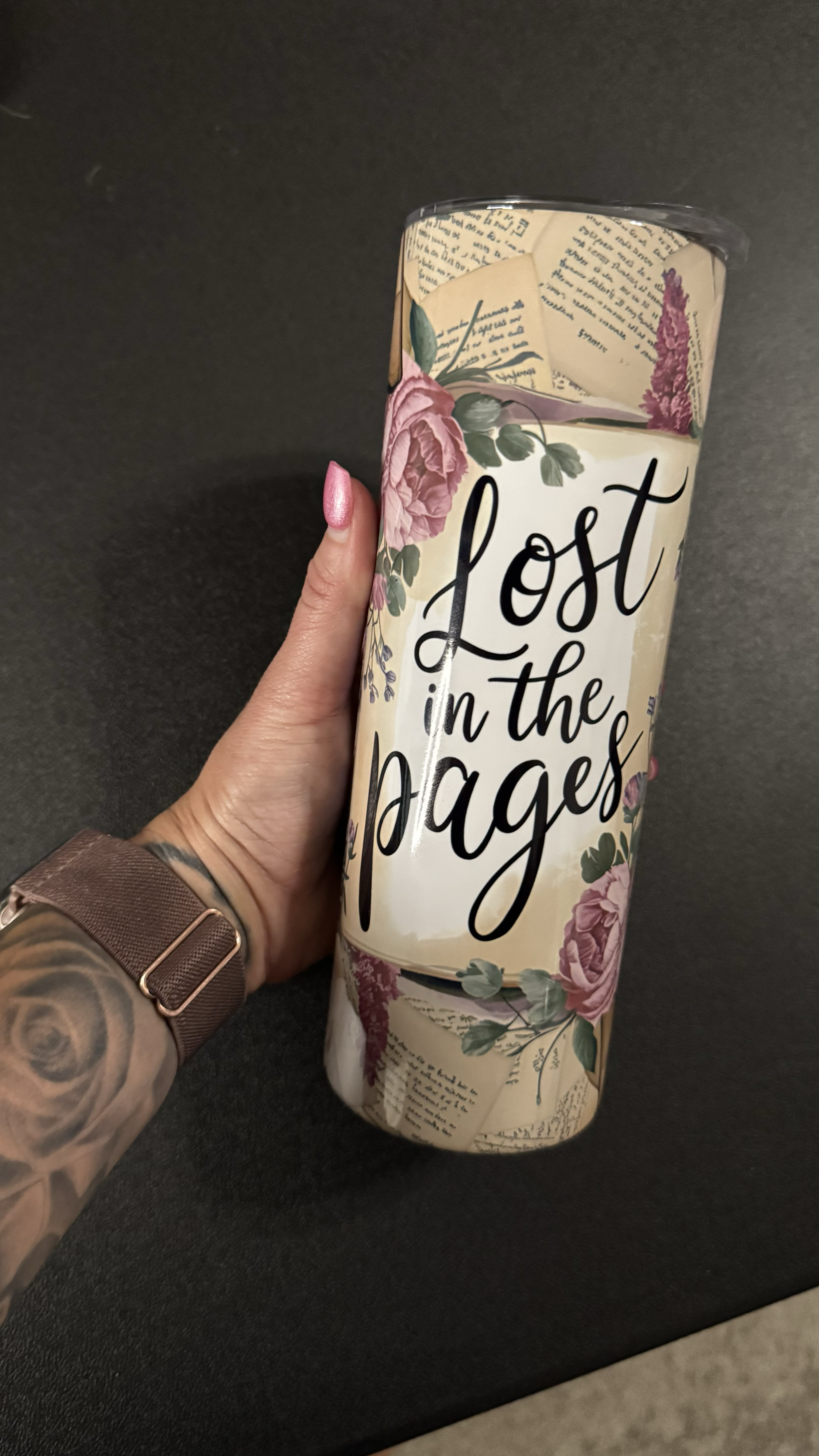 A hand holding a cylindrical floral-themed container with the text "Lost in the pages" on it, and a wristwatch on the wrist.
