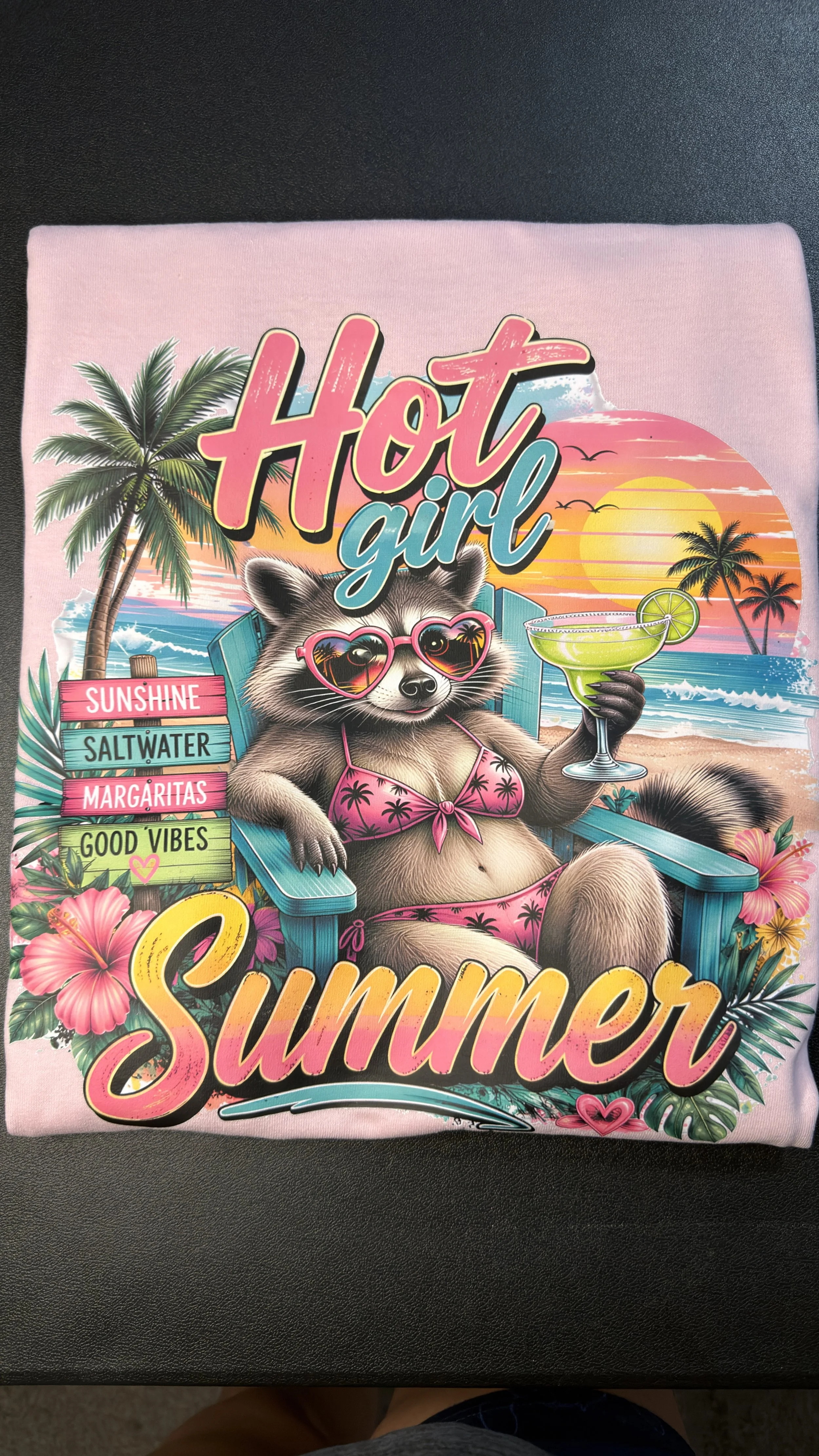Colorful illustration of a raccoon wearing sunglasses and a bikini, sitting on a beach chair holding a margarita, with a sunset, palm trees, and ocean background. The text says 'Hot Girl Summer' and includes words like 'Sunshine,' 'Saltwater,' 'Marga