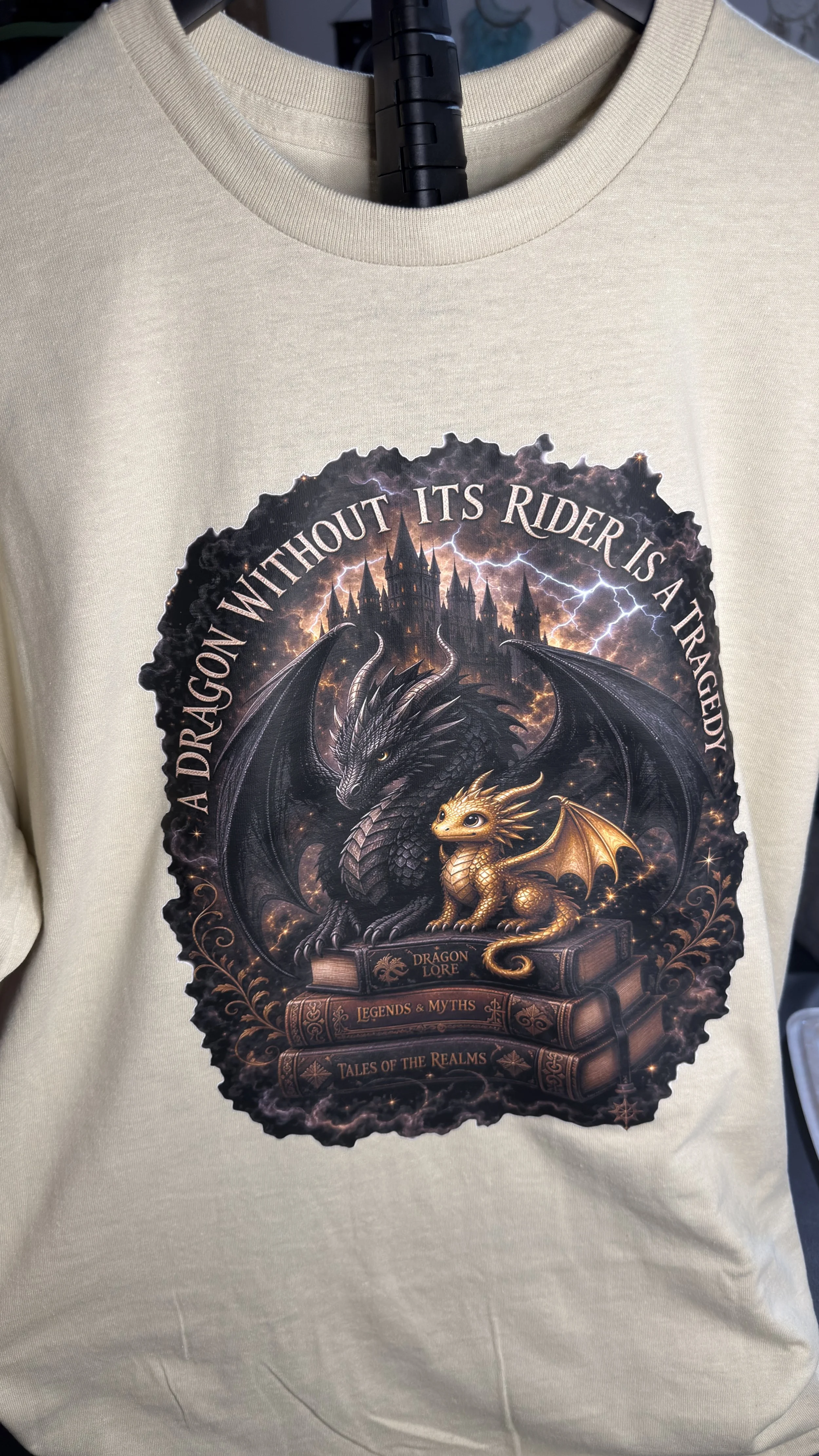 A white shirt displaying a graphic design of a black dragon and a small golden dragon, with a background of a dark castle and lightning. The text says, "A Dragon Without Its Rider Is a Tragedy."