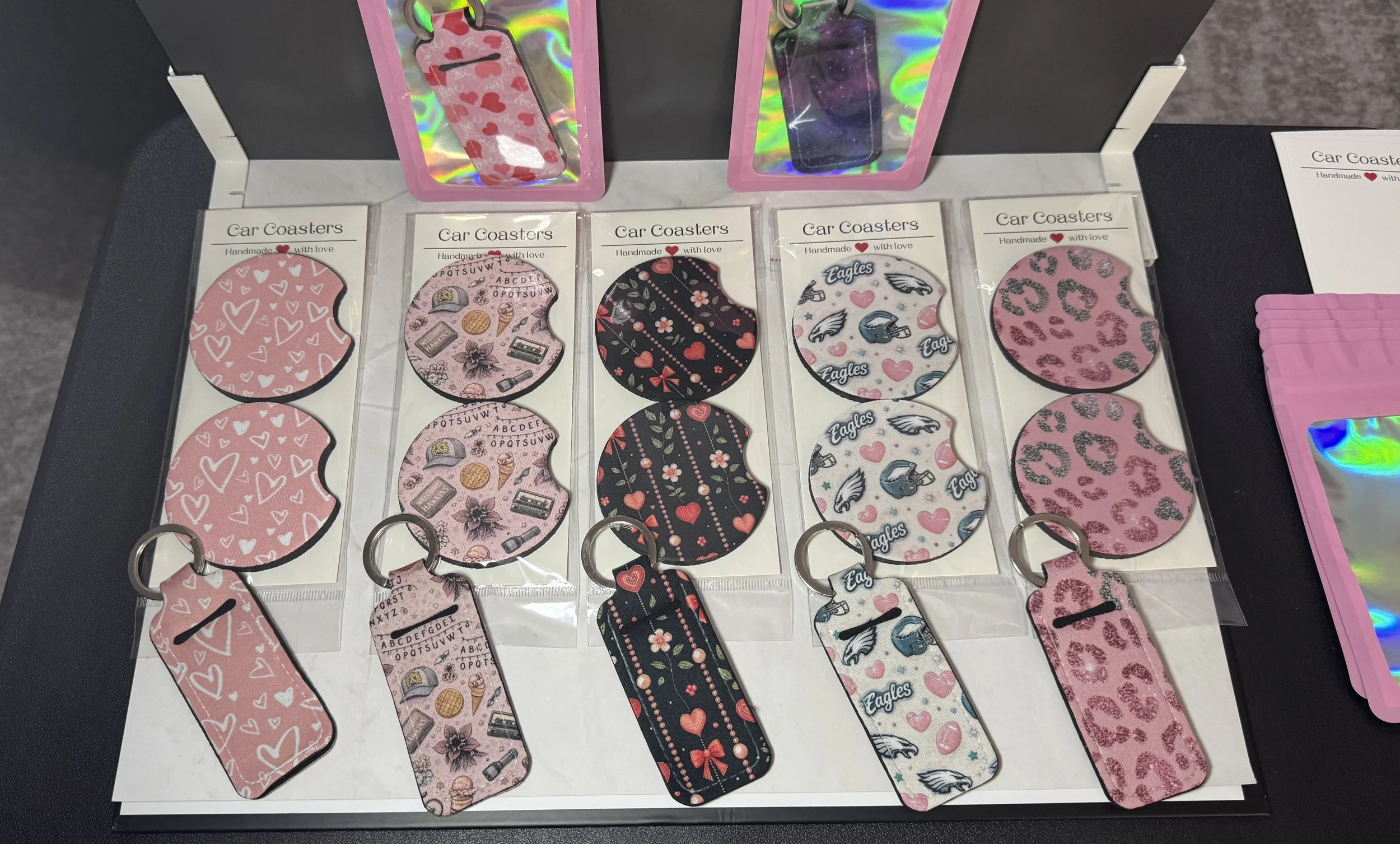 Display of colorful keychains shaped like car coasters with various patterns, including hearts, gifts, sports teams, and animals, attached to metal rings and arranged in rows on a white tray.