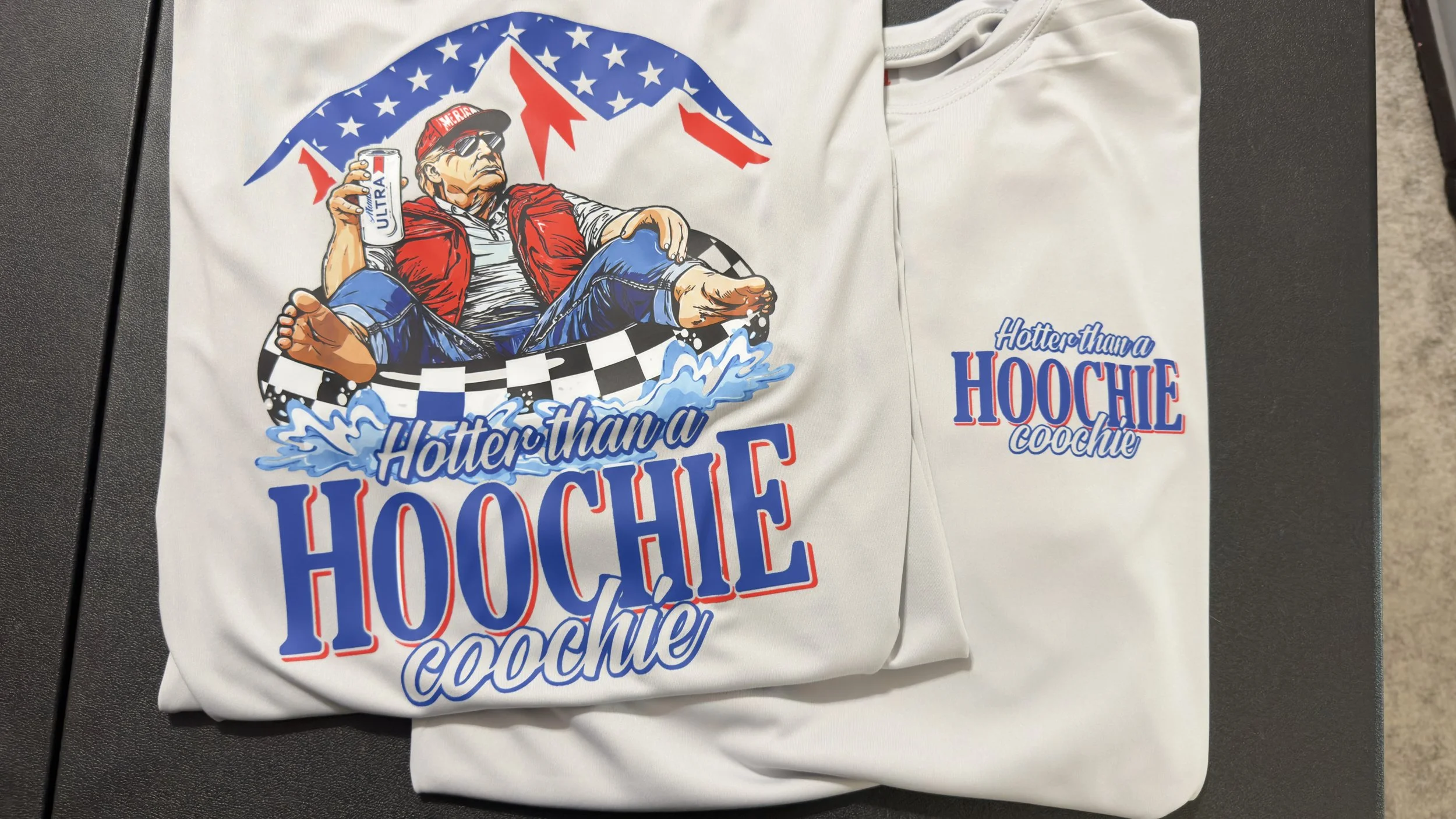 A white t-shirt with a graphic of an older man wearing sunglasses, a red vest, and a baseball cap holding a can of Ultra beer, sitting in a boat in water, with the text "Hotter than a Hoochie Coochie" in red and blue.