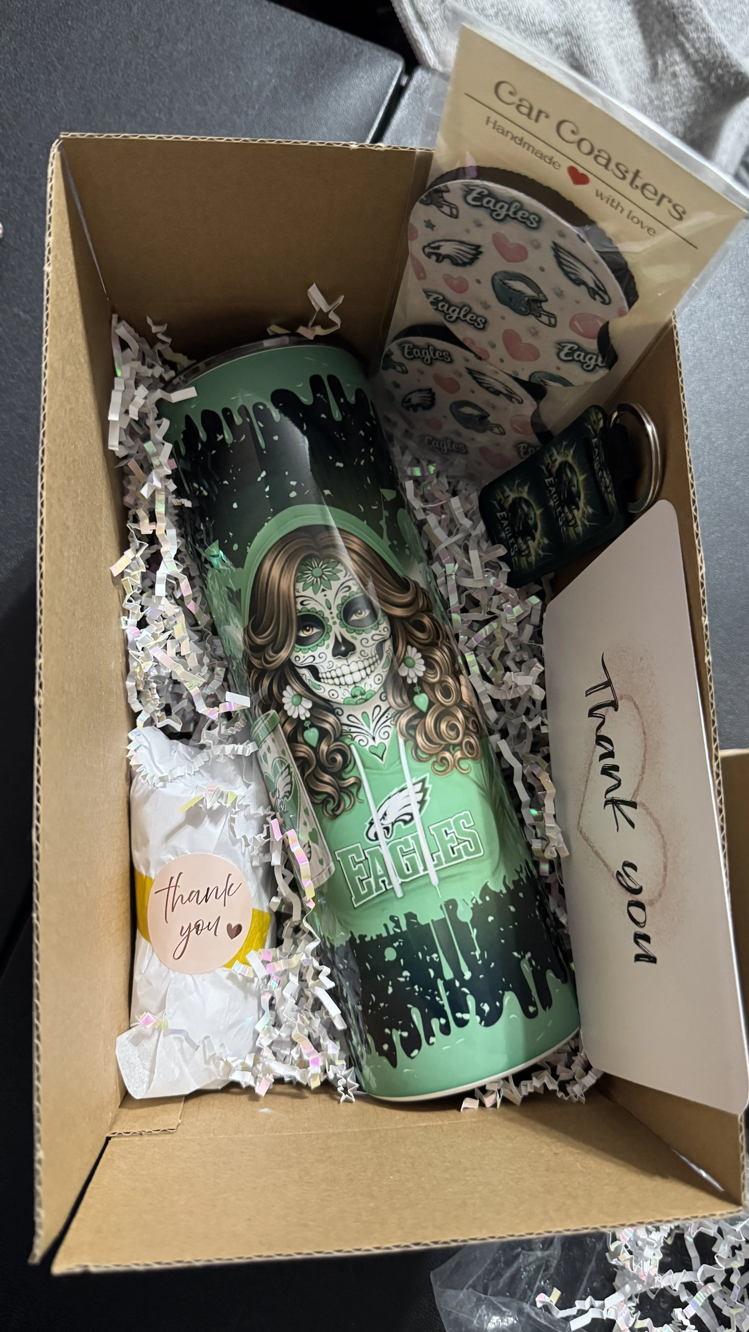 A gift box containing a Eagles-themed tumbler with a Day of the Dead style skull design, decorative paper shreds, Eagles packaging, a thank you note, and a small bottle of Eagles-themed shot.