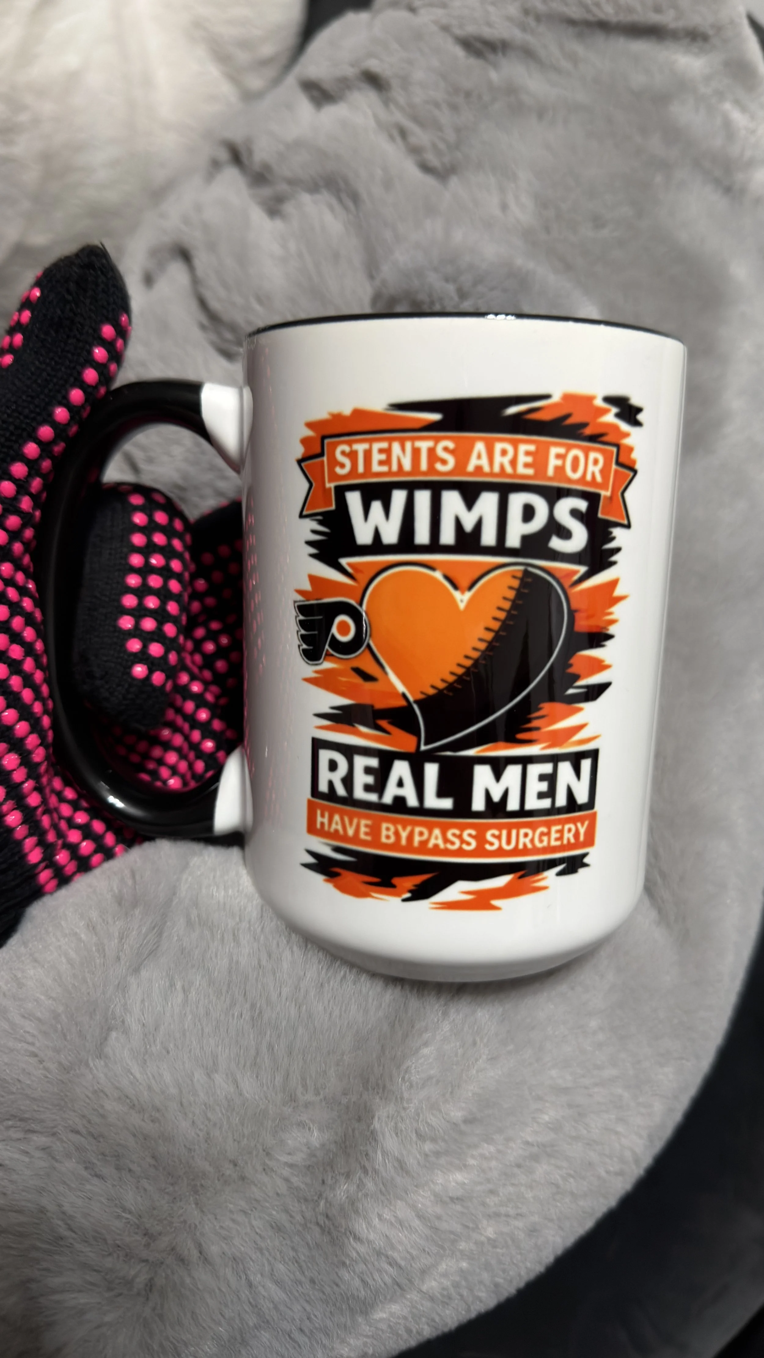 A white mug with bold text that reads, "Stents are for Wimps, Real Men Have Bypass Surgery." The mug features a heart graphic with a stent and surgical tools. It is held by a hand wearing a black glove with pink dots, and the background appears to be