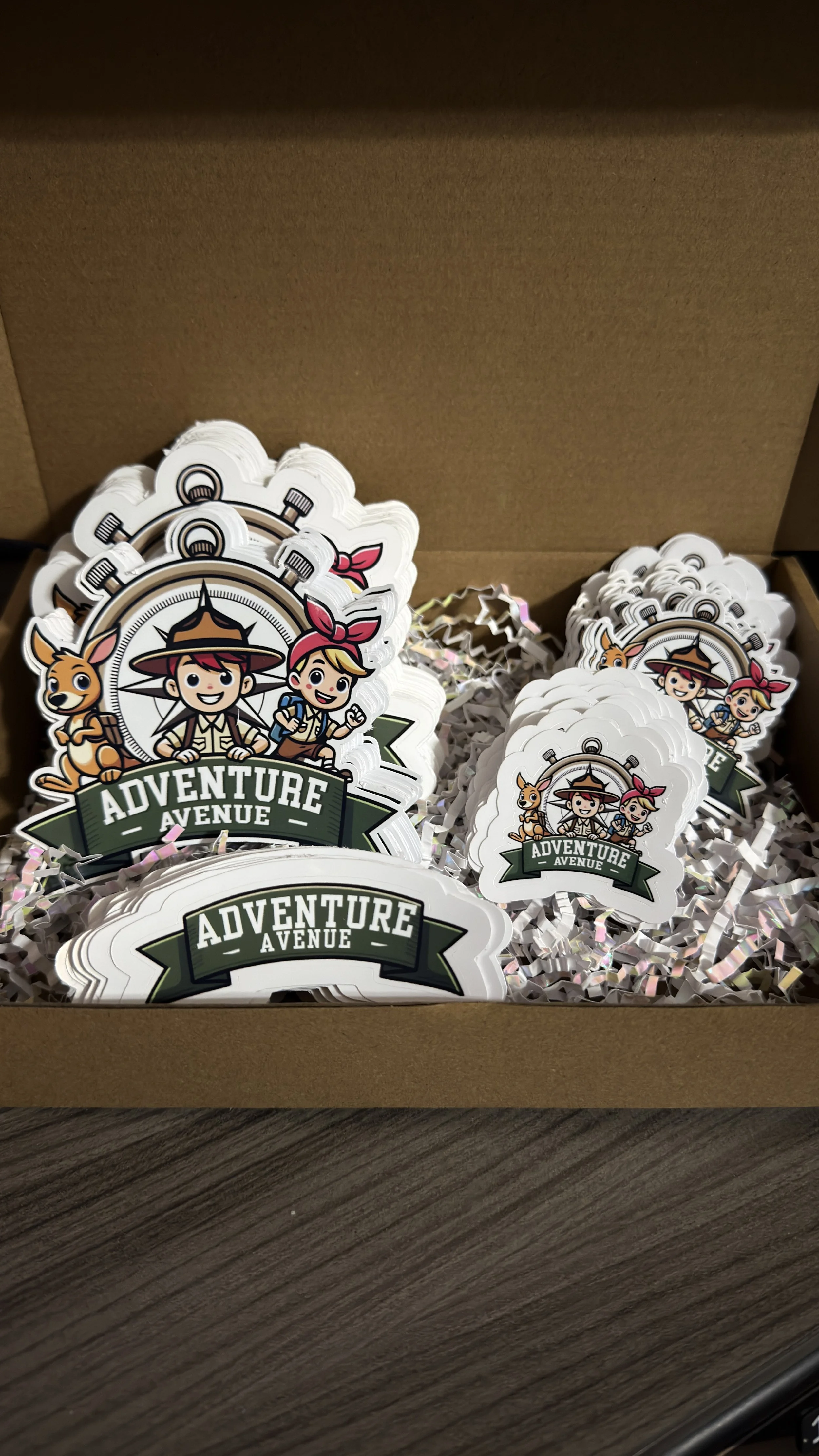 A box of stickers featuring cartoon characters of a boy with a hat, a girl with a pink bow, and a dog, with the text 'Adventure Avenue' on a green banner.