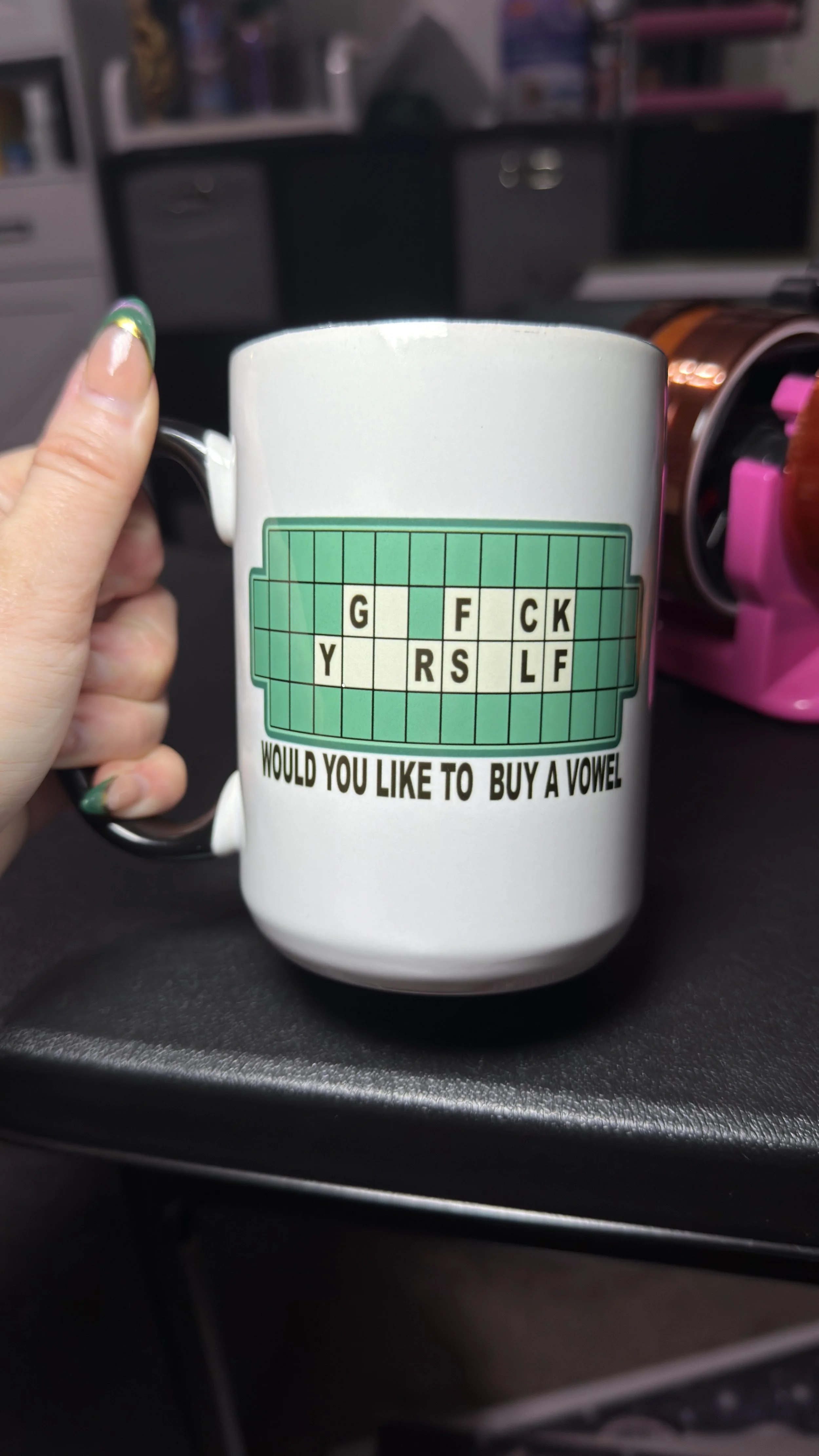 White coffee mug with a green crossword puzzle and the text "Would you like to buy a vowel" printed on it, held by a person with painted nails, with a blurred background of office supplies.