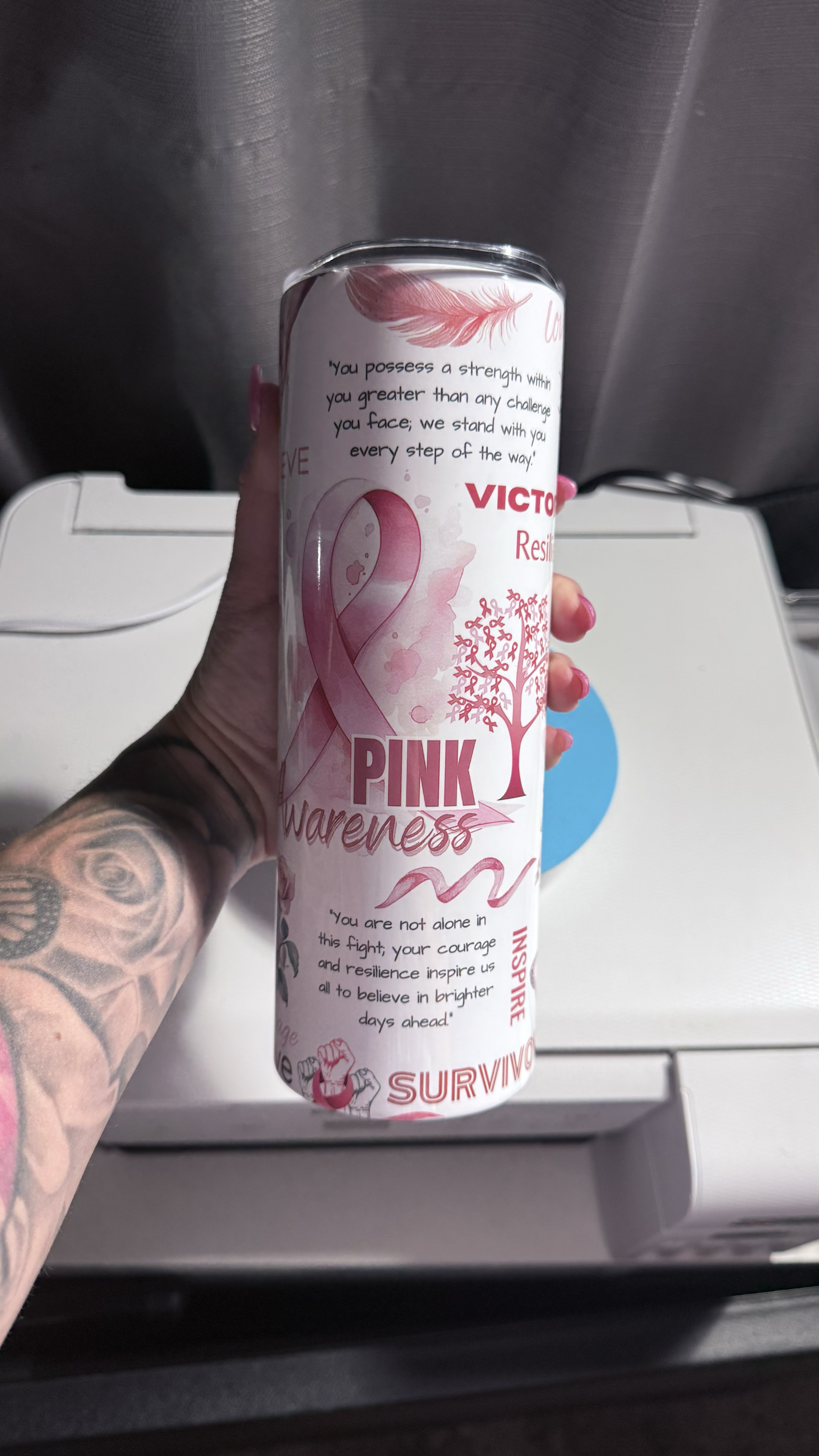 A hand with tattoos holding a pink ribbon awareness can on an airplane tray table. The can promotes breast cancer awareness and resilience.