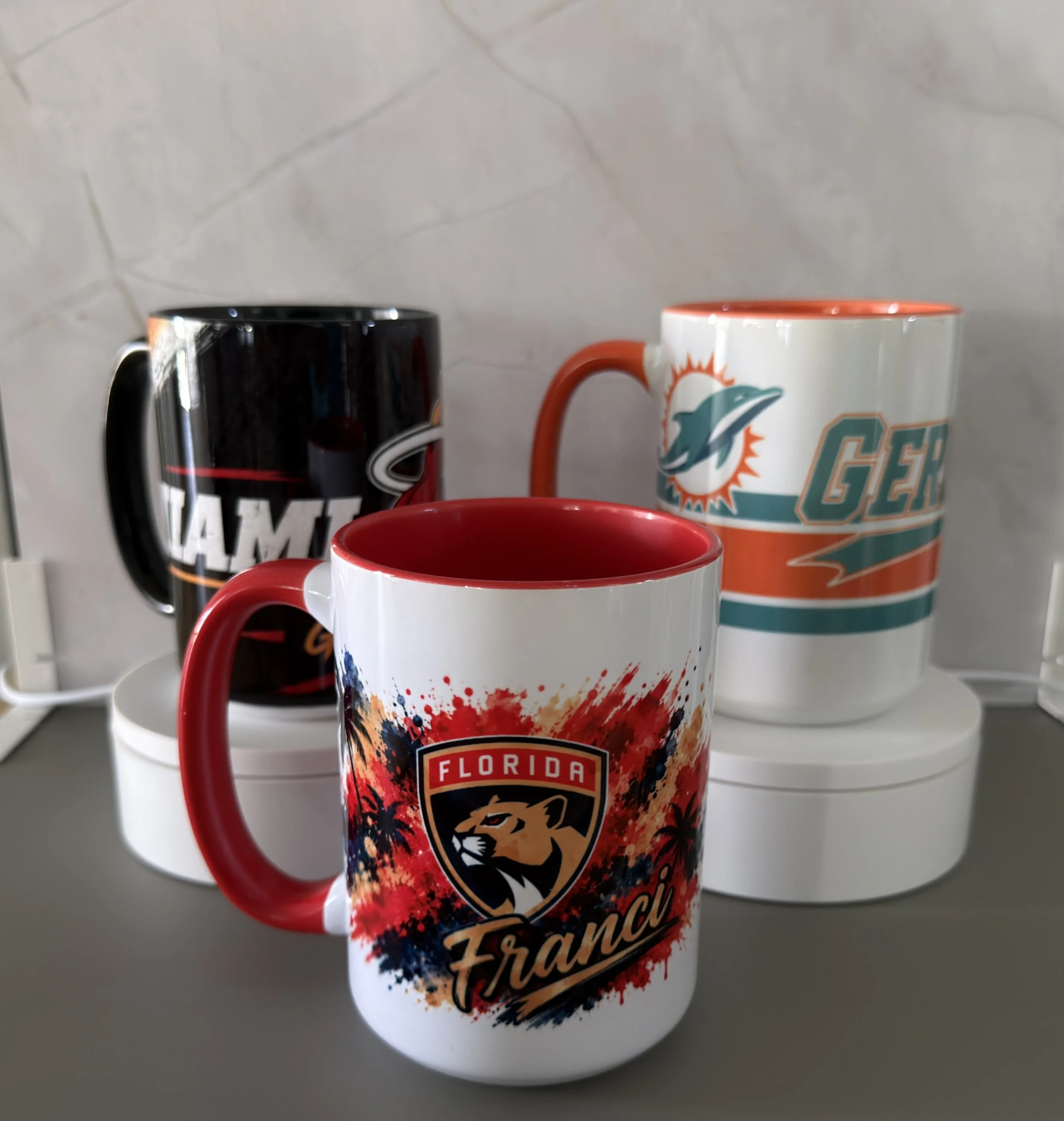 White ceramic mug featuring the Miami Dolphins logo with an orange handle, and a black ceramic mug with a logo design, both placed on a white shelf against a marble wall.