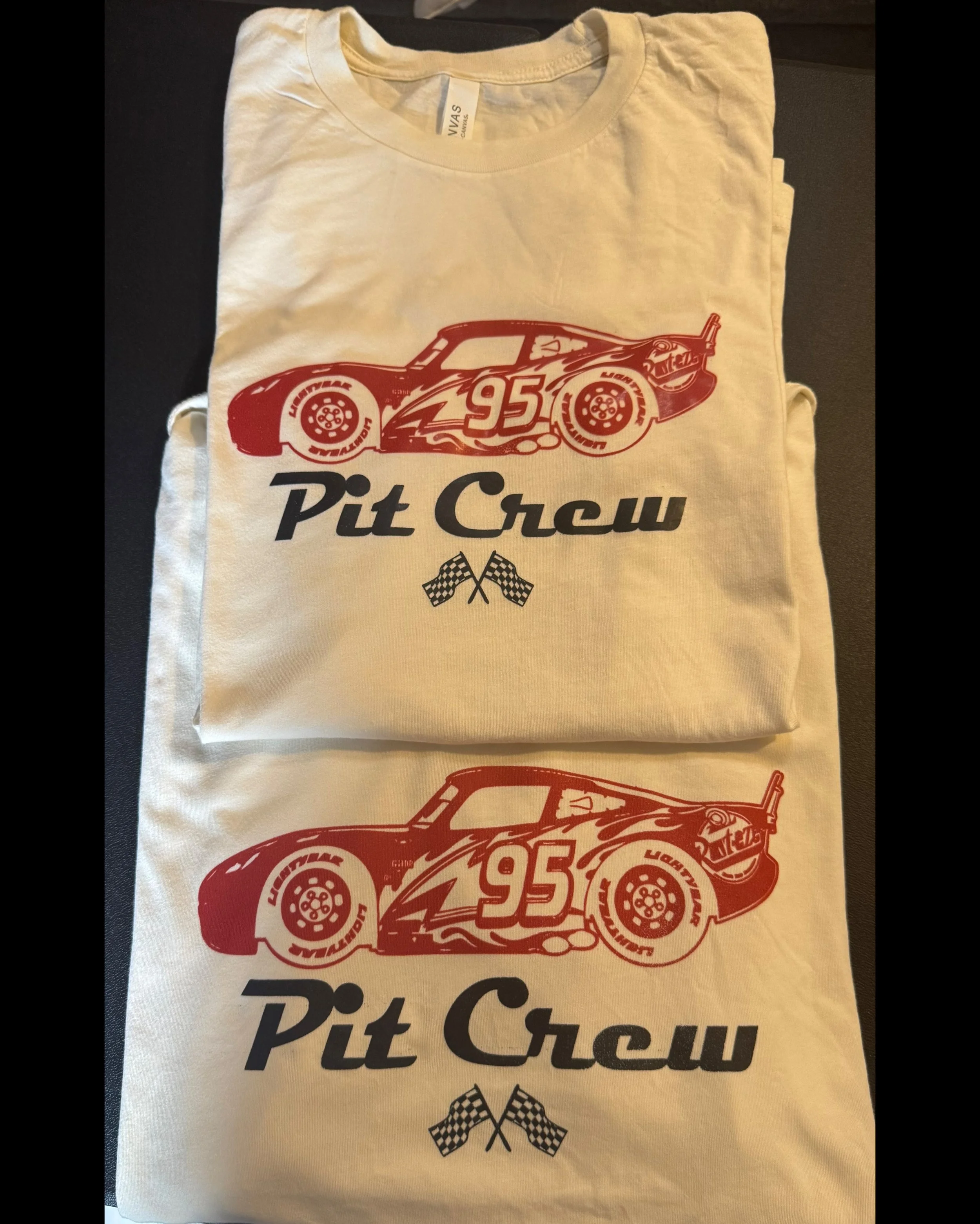 A white T-shirt with a graphic design of a race car and the number 95, along with the text "Pit Crew" and crossed checkered flags underneath.