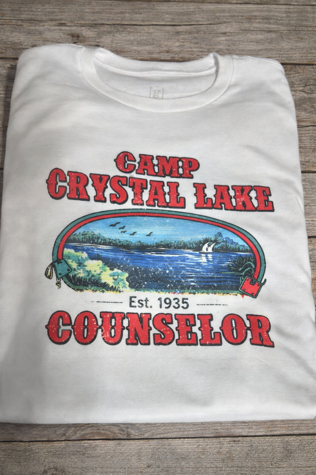 White T-shirt with a colorful lakeside scene and text that reads "Camp Crystal Lake," "Est. 1935," and "Counselor."