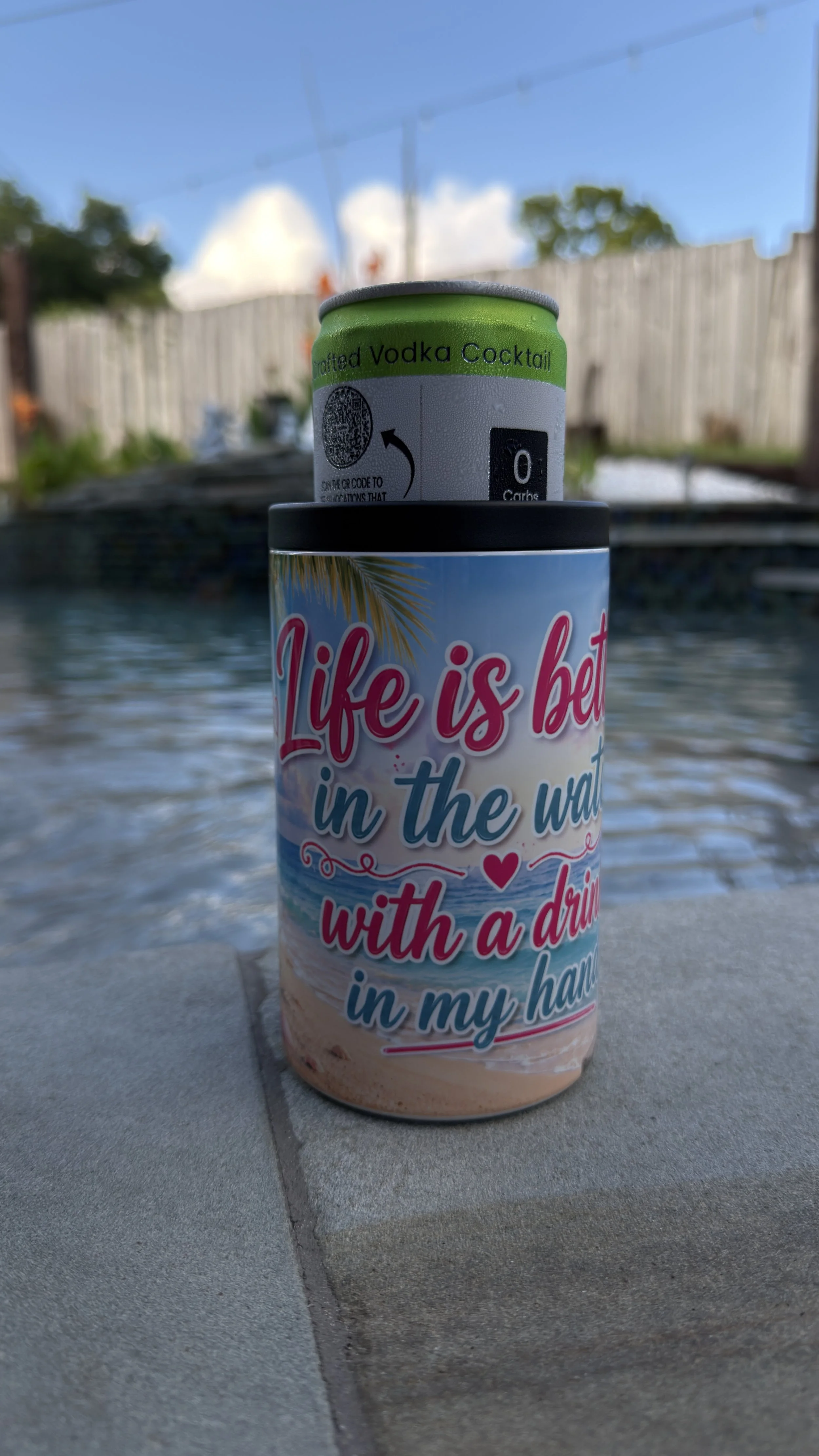 Close-up of a colorful drink can with a beach scene and the words "Life is better in the water with a drink in my hand" written on it, placed on a poolside surface.