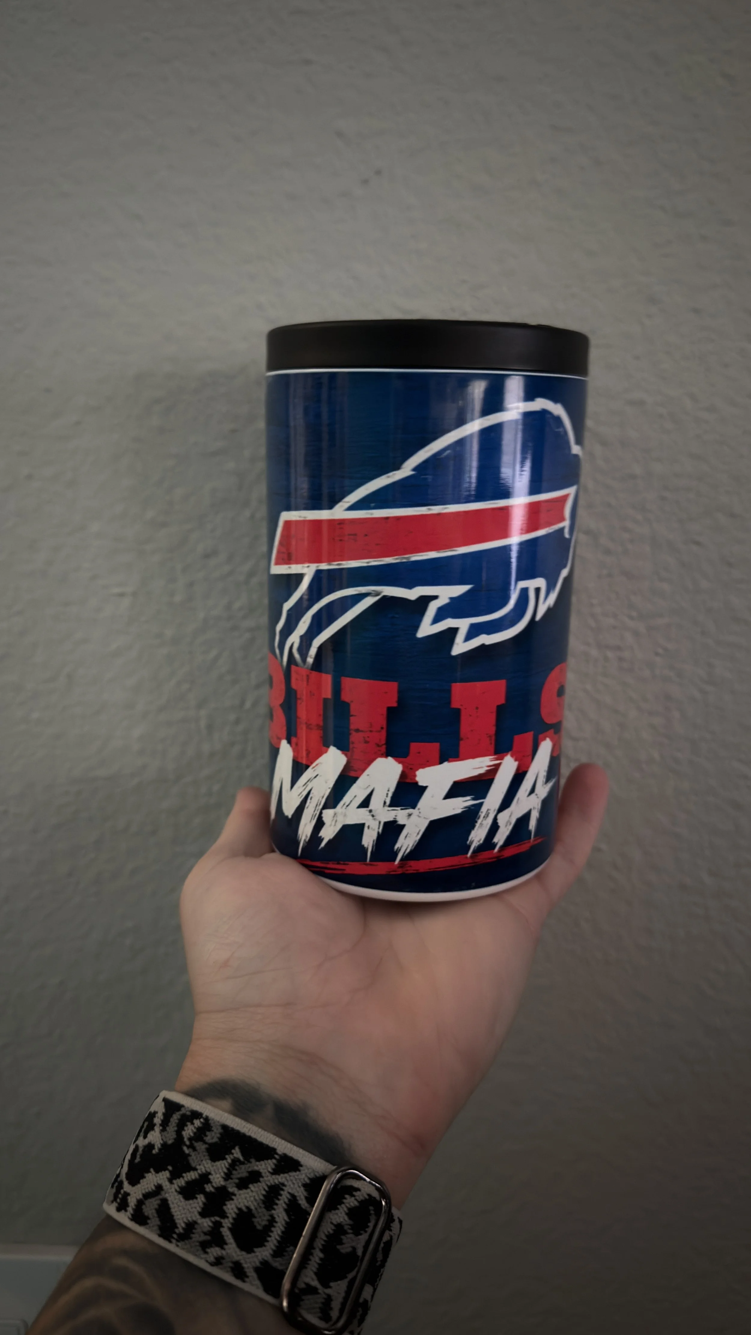 A hand holding a cylindrical container with a Buffalo Bills logo and the text 'BILLS MAFIA' against a plain textured wall background.